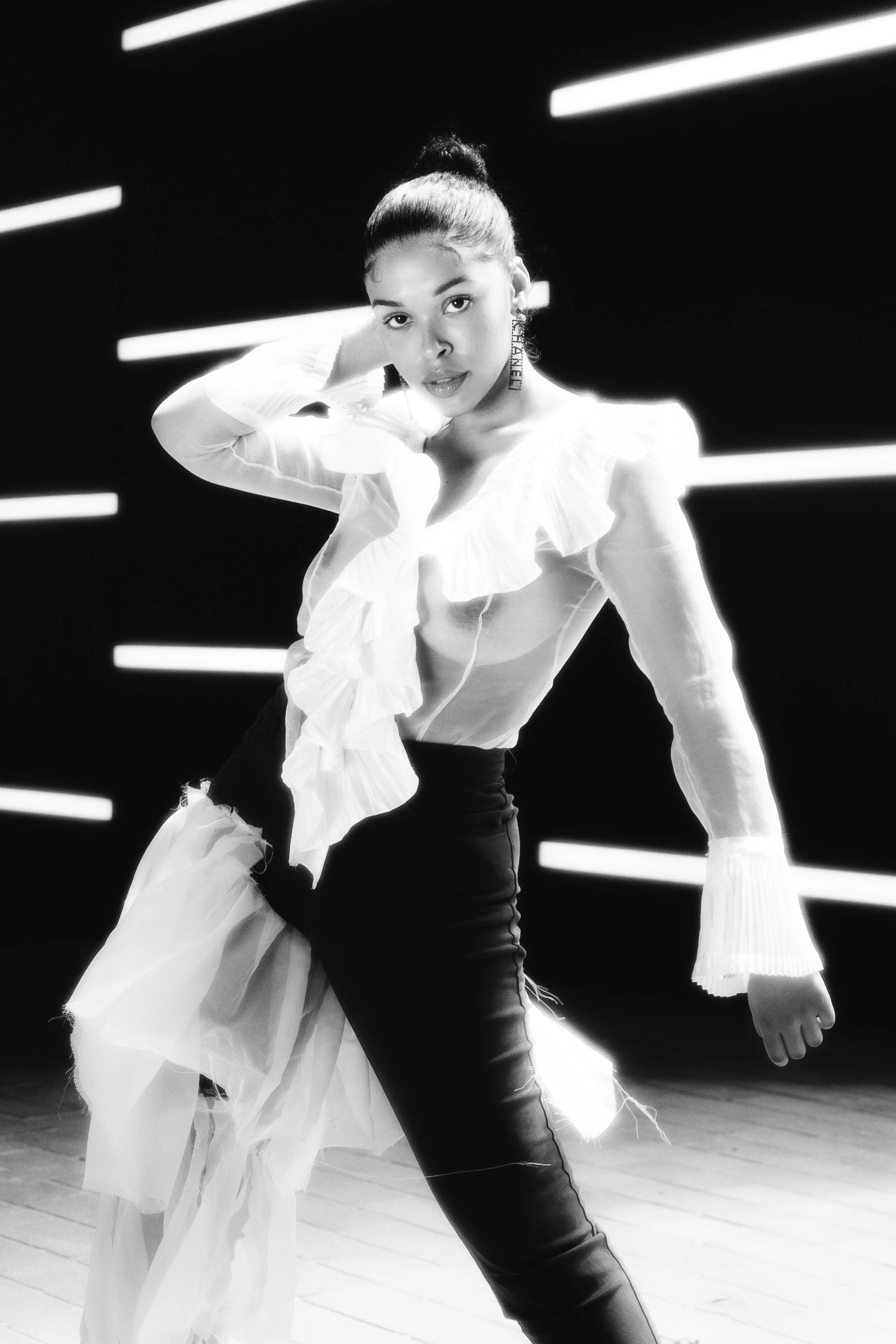 A woman posing on a stage with a dark background and horizontal light bars. She is wearing a sheer white blouse with ruffles and black pants, with her hair in a bun.