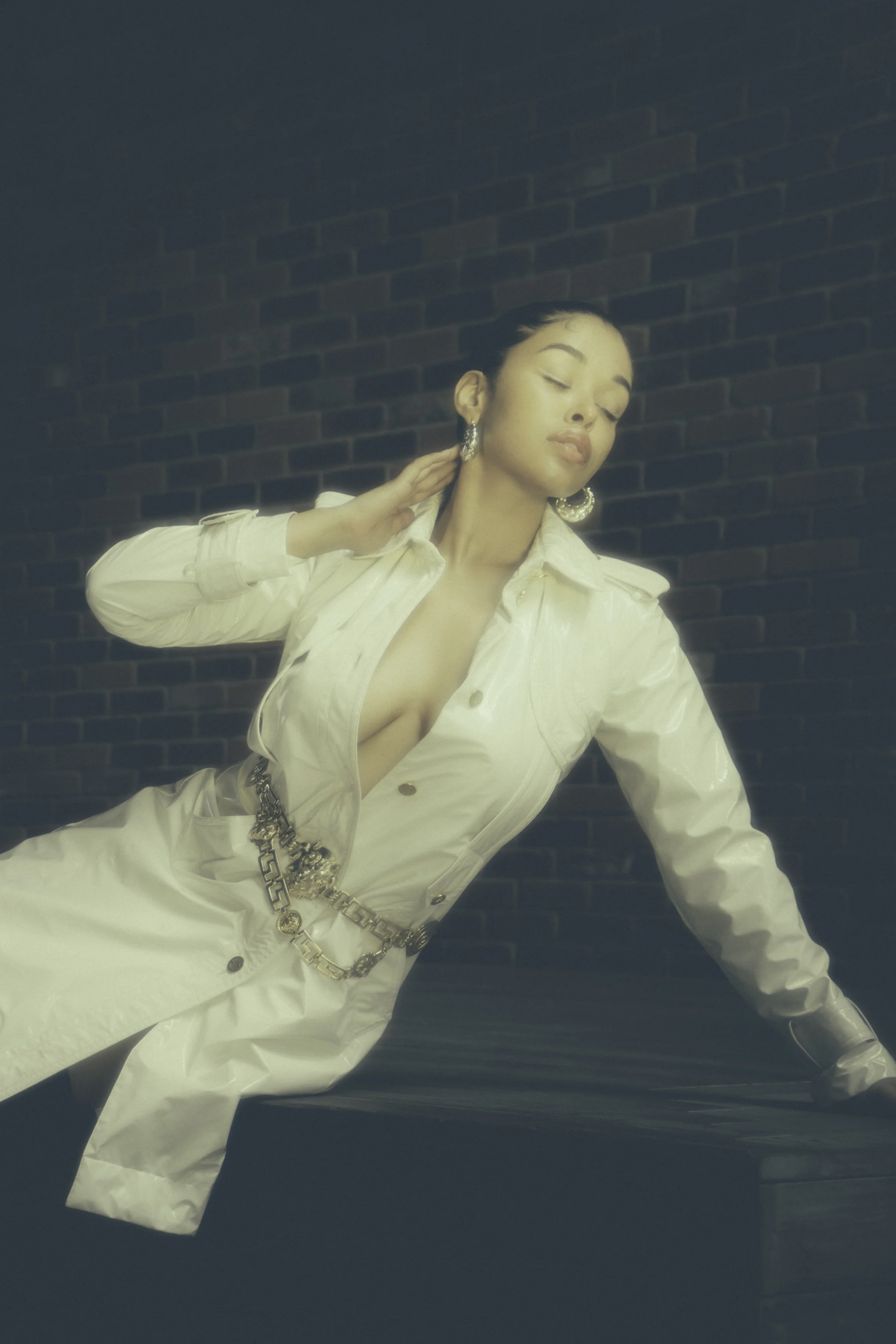 A woman in a cream-colored trench coat with a deep neckline, wearing large earrings and a chain belt, poses against a dark brick wall.