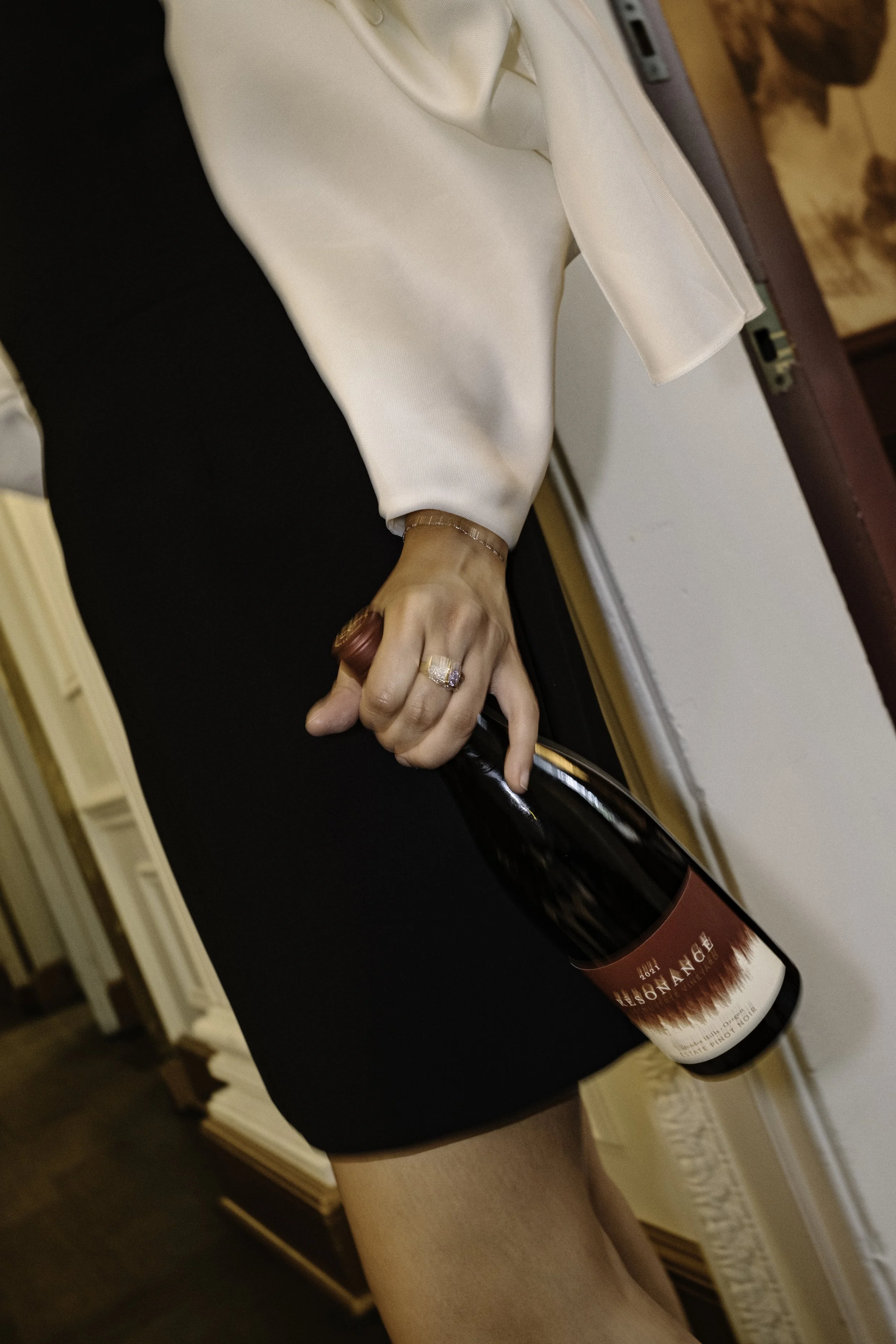 A woman dressed in formal attire holding a bottle of wine in her right hand, with her lap and part of her arm visible.