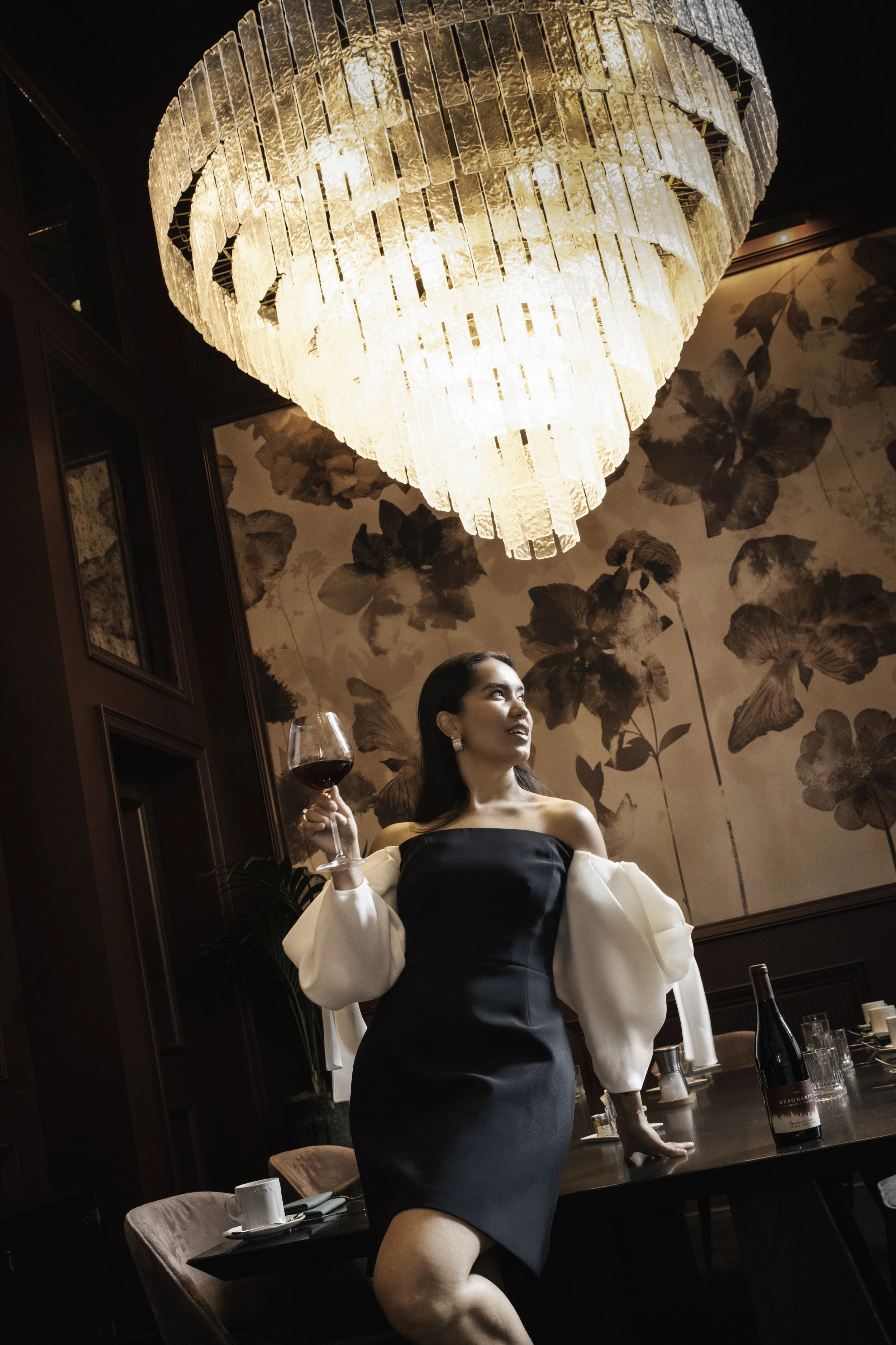 A woman in a black off-shoulder dress with puffy white sleeves holding a glass of red wine in a restaurant with dark wood paneling and floral wallpaper, under a large, elegant chandelier.