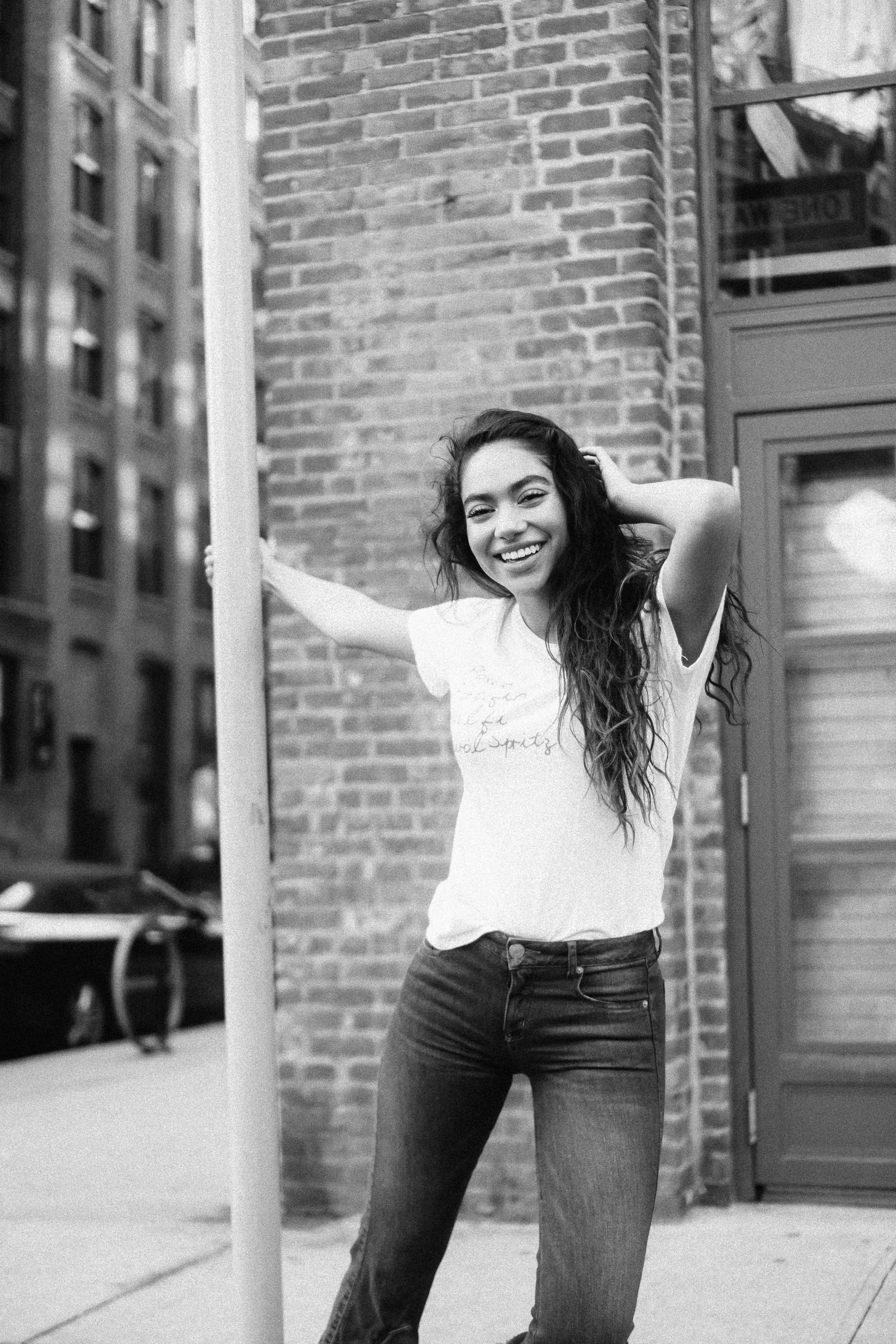 A young woman with long, wavy hair smiling, standing outdoors next to a brick building with a storefront.