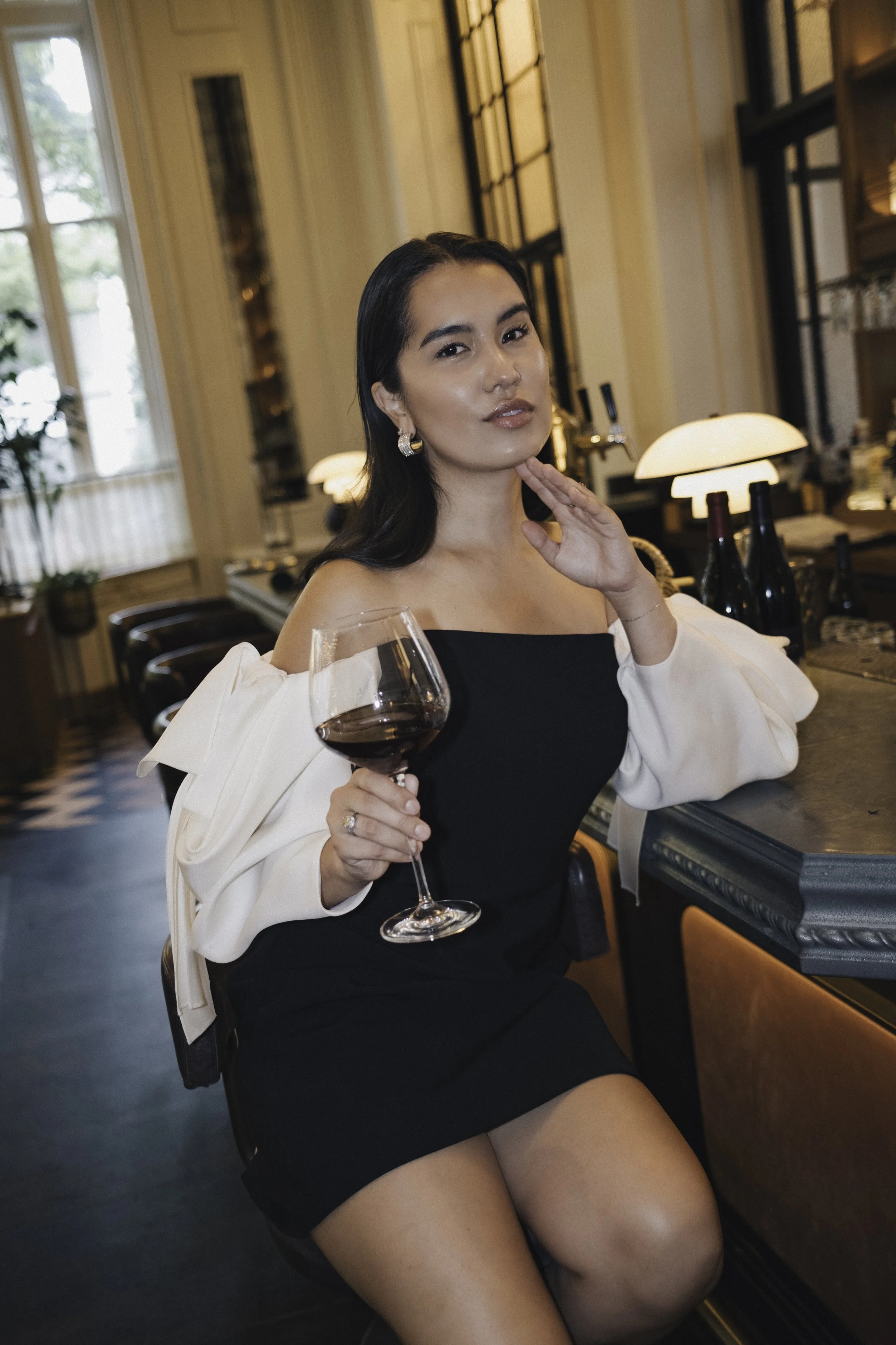 A woman sitting at a bar holding a glass of red wine, wearing a black off-the-shoulder dress and white jacket in a sophisticated restaurant or bar with warm lighting and decorative interior.