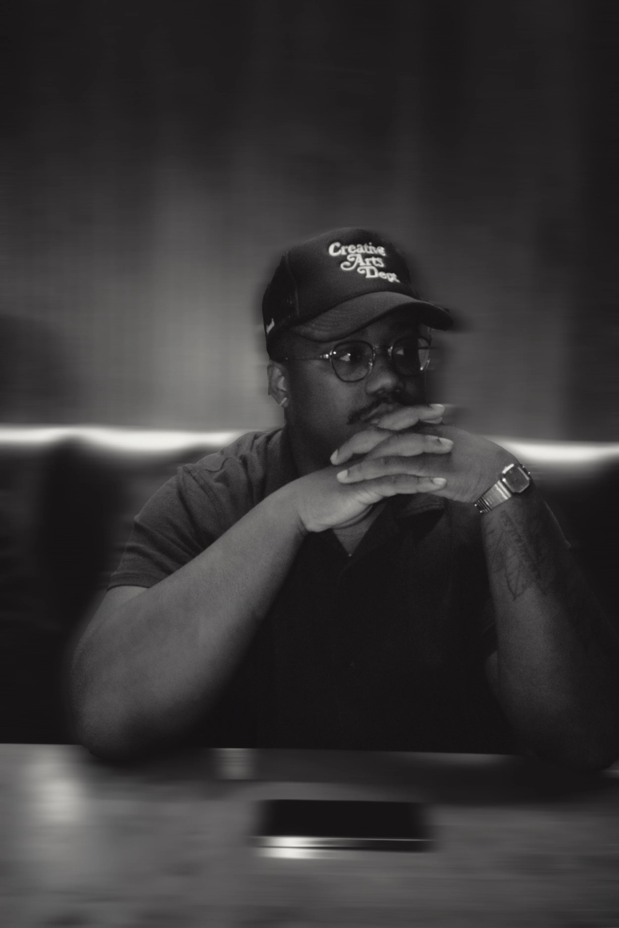 A man wearing glasses and a cap that reads "Creative Arts Dept" is seated at a table with his hands clasped and resting near his face, in a black and white photo.