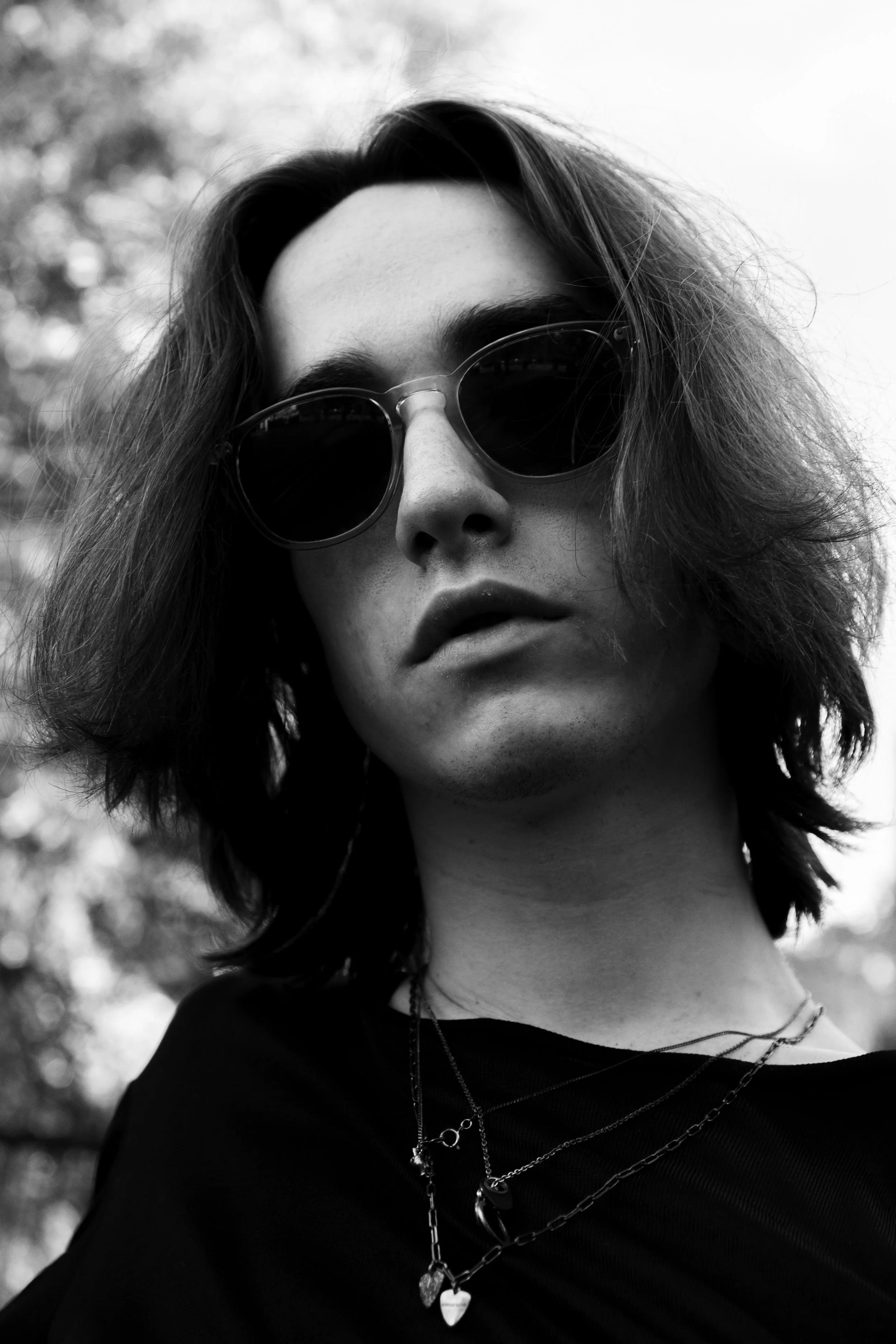 A black and white close-up photo of a woman with medium-length wavy hair, wearing sunglasses and layered necklaces, looking slightly upwards.