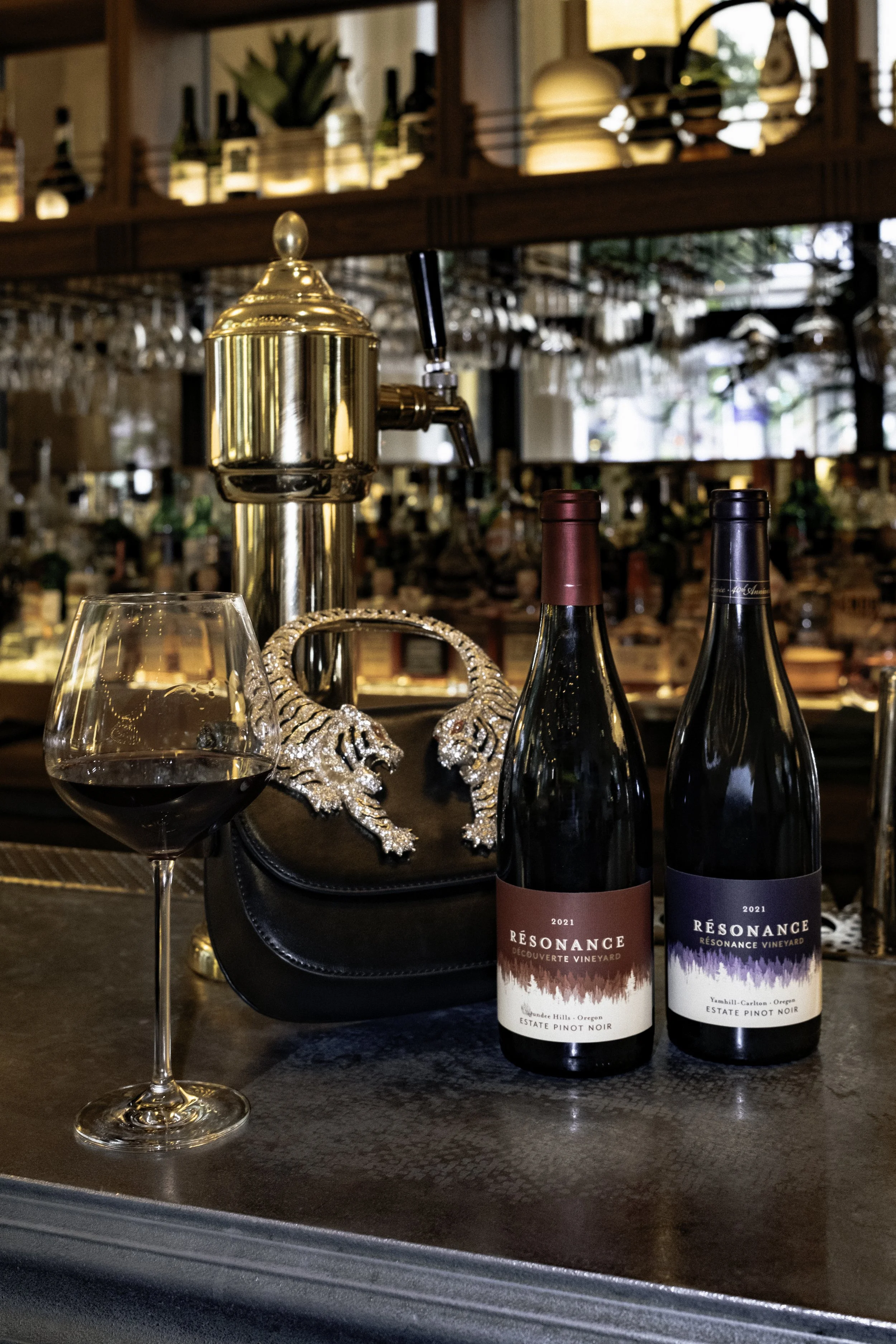 A glass of red wine, two bottles of wine, a black purse with silver snakehead decoration, and a gold wine tap on a bar counter.