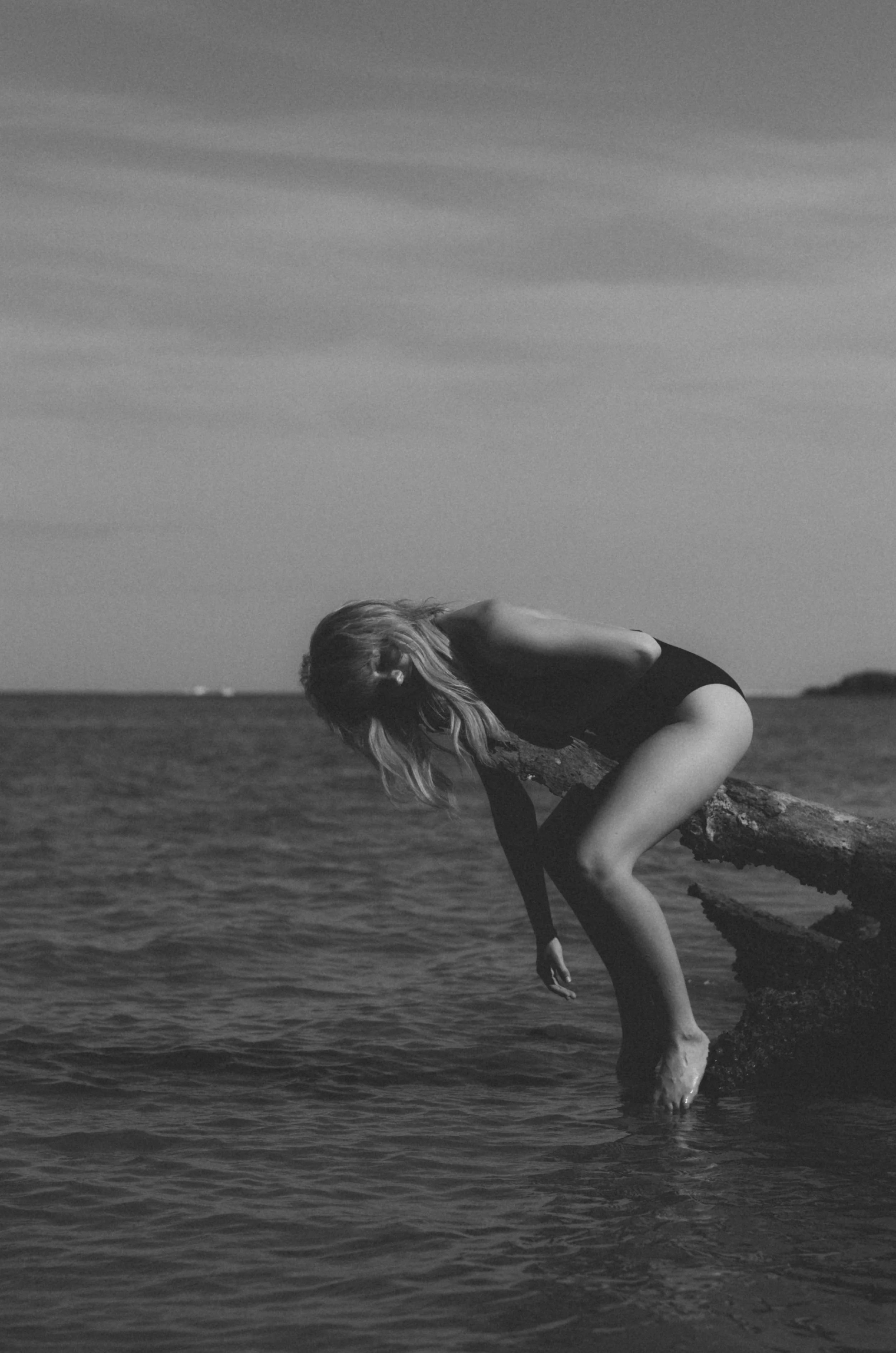 A woman in a black swimsuit leaning over a rock on the shoreline with her feet in the water, looking down at the water, in black and white.