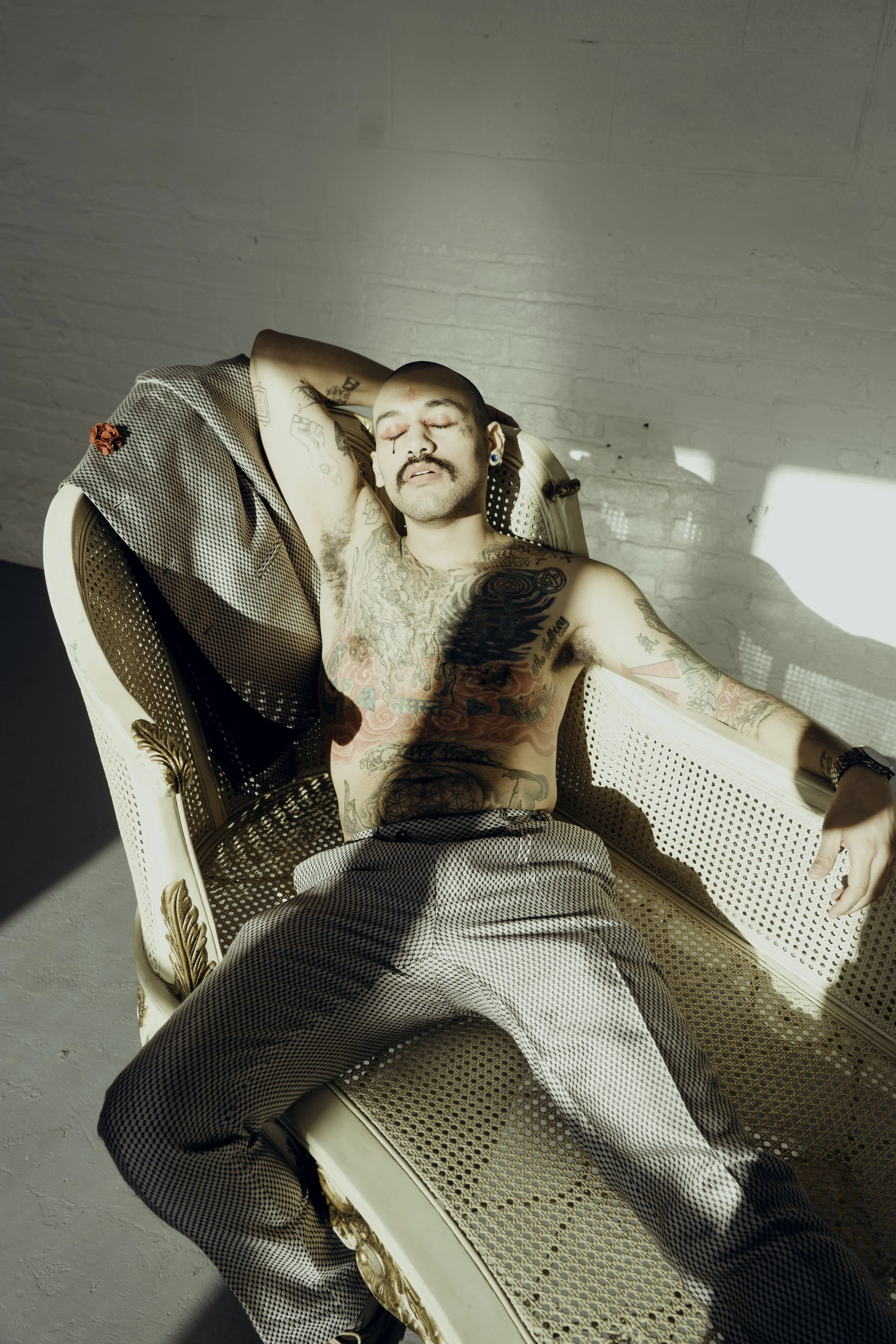 A shirtless man with tattoos, a mustache, and earrings lounging on a vintage-style cream-colored sofa with gold accents, with sunlight casting shadows on a white brick wall.