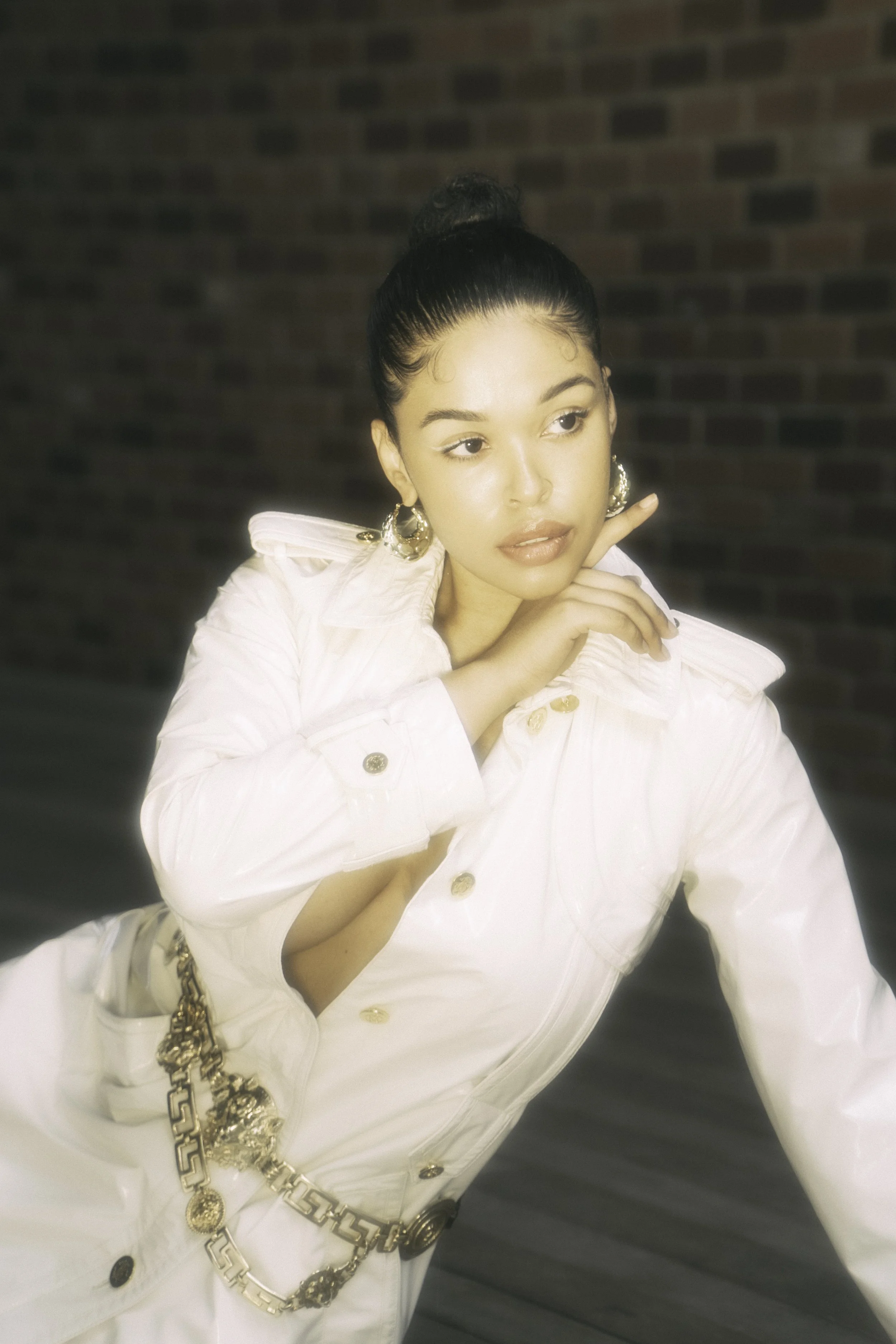 A woman with dark hair in a high bun, wearing large gold hoop earrings and a white jacket with gold buttons and chain accessories, stands against a brick wall background, posing with her hand near her face and looking to the side.