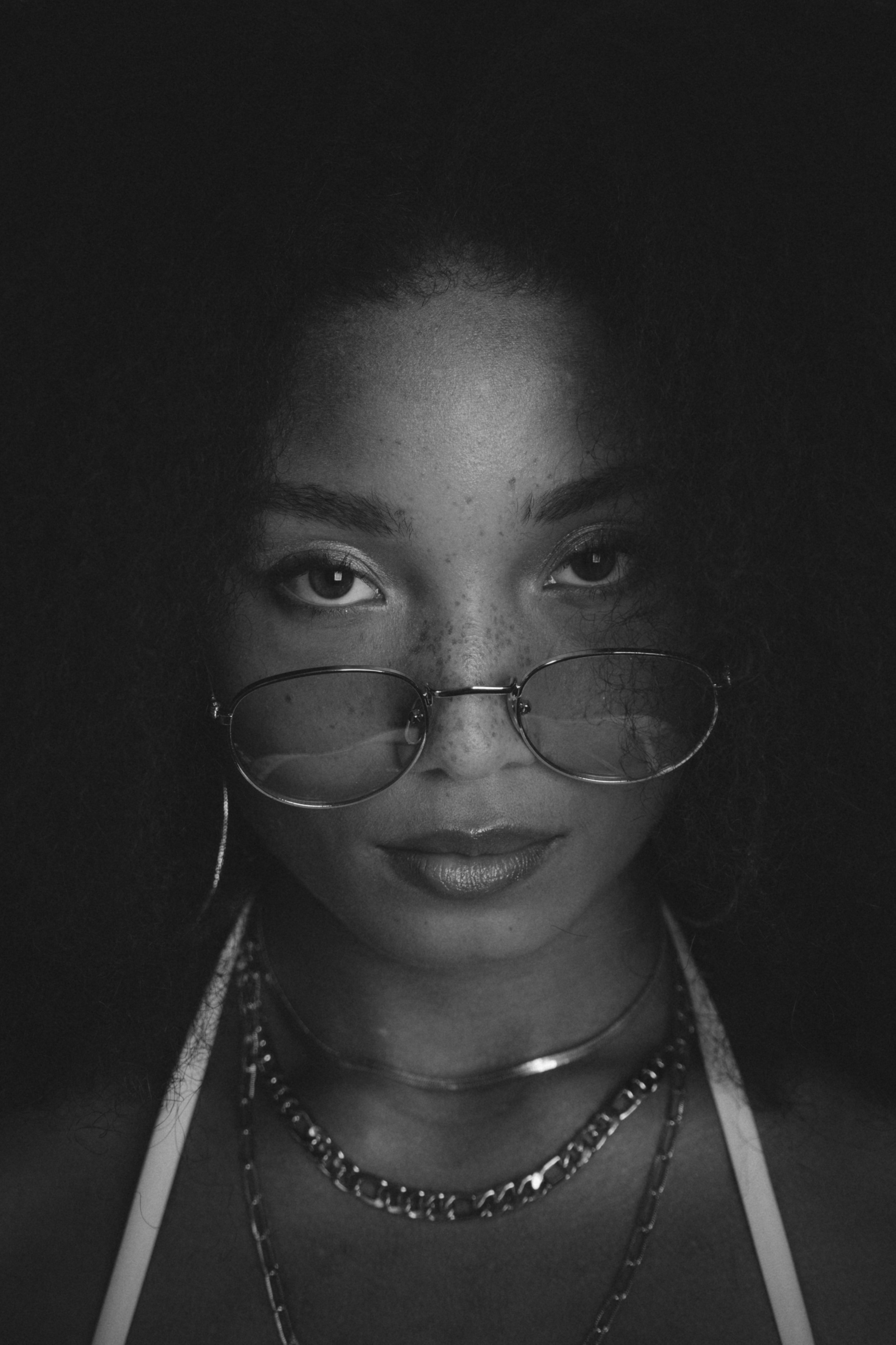 Black and white portrait of a woman with curly hair, wearing glasses, hoop earrings, necklaces, and a tank top.