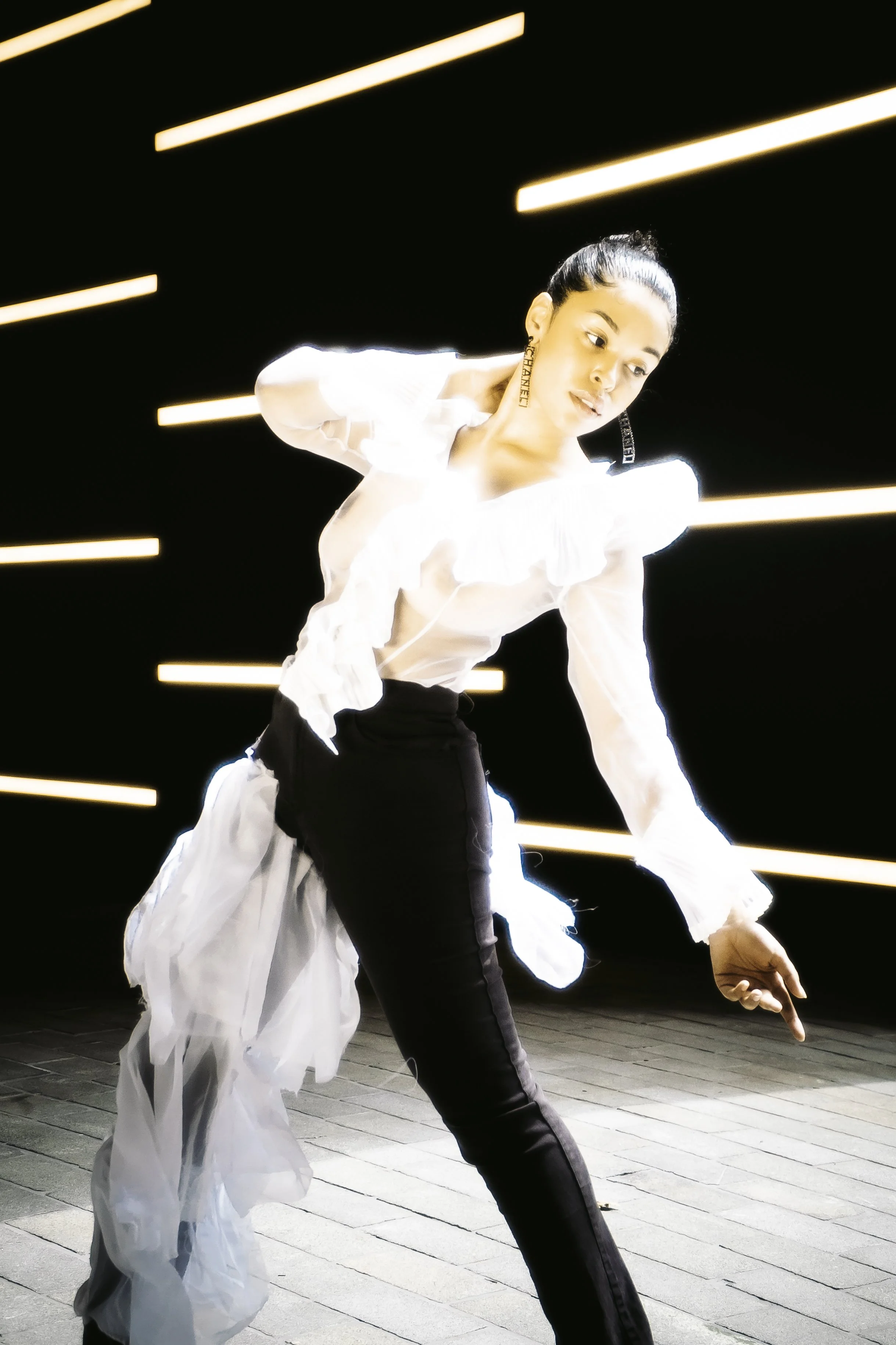 A woman in a white ruffled blouse and black pants poses against a black background with yellow horizontal light bars, creating a striking visual effect.