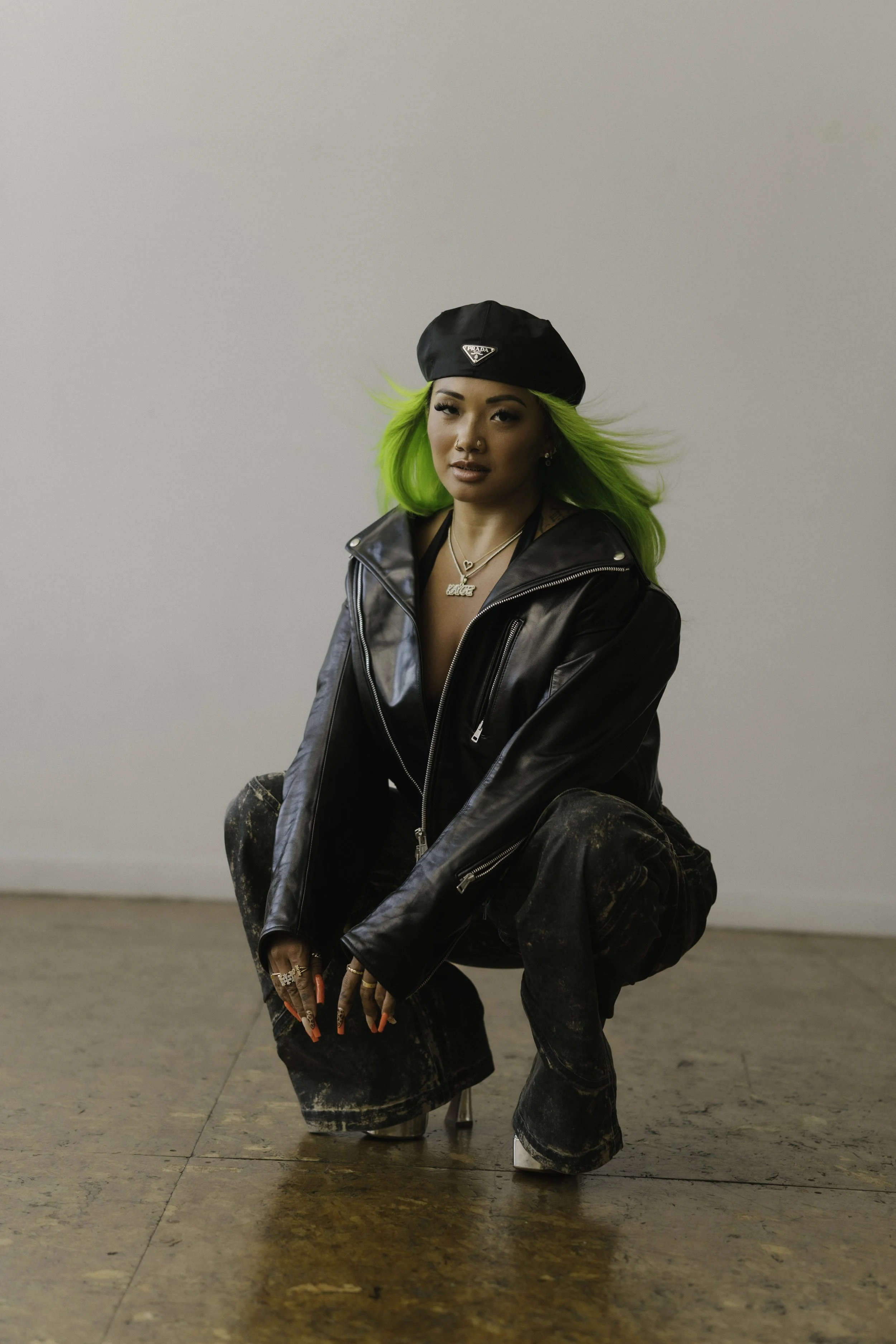 A woman with bright green hair wearing a black beret, a black leather jacket, and black distressed pants, squatting in a room with a plain white wall and wooden floor.