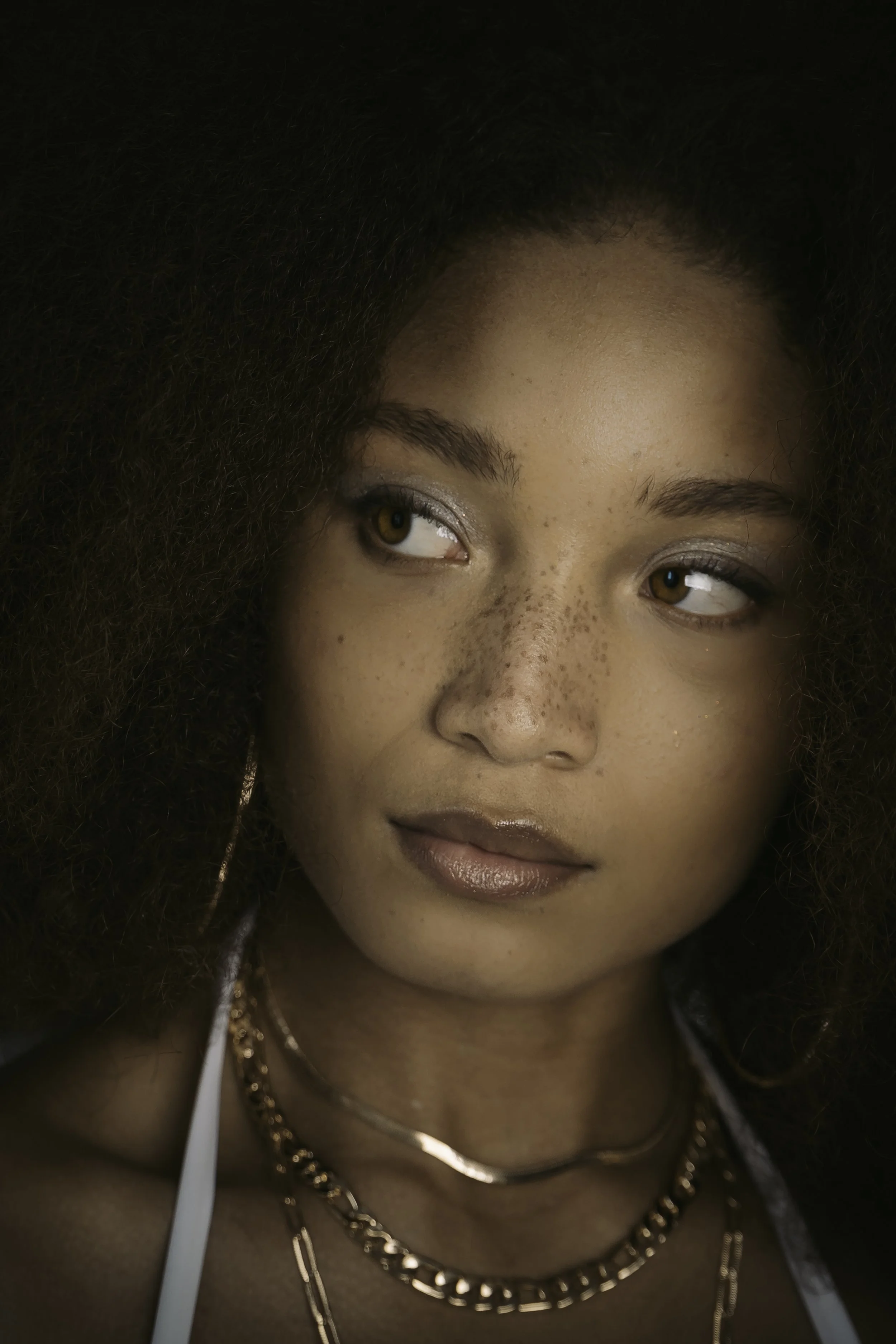 Close-up of a woman with curly hair, light makeup, and gold jewelry, looking to the side with a neutral expression.