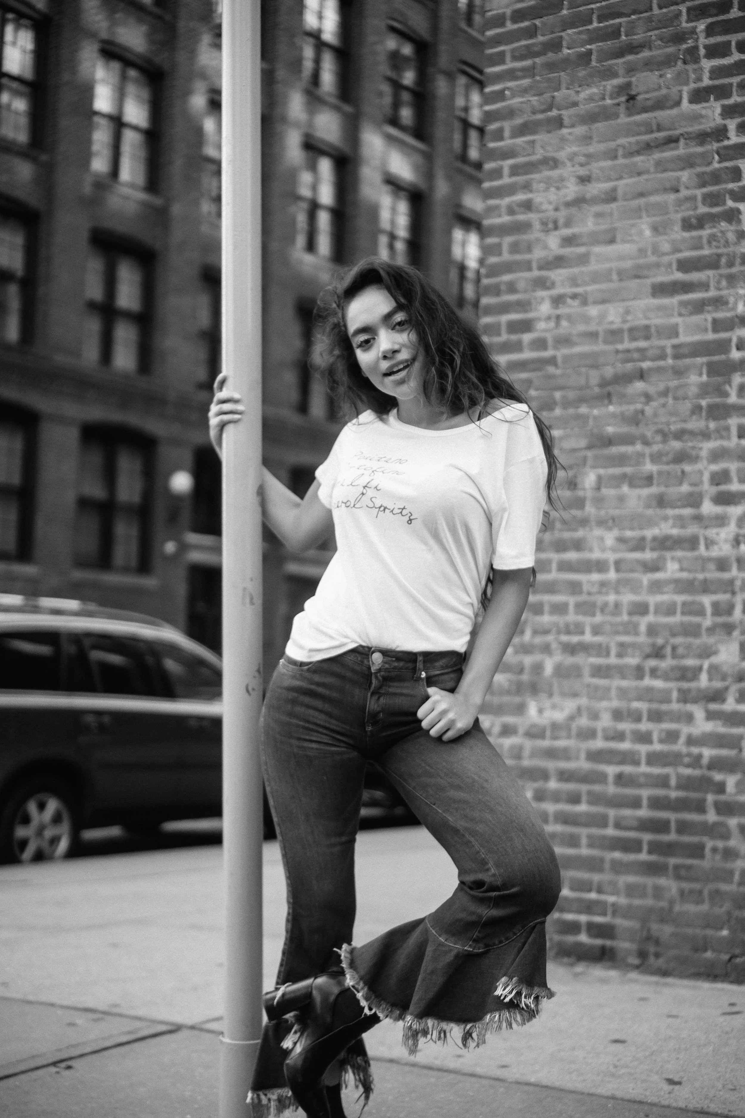 A woman standing on a city sidewalk, leaning on a pole with one hand, wearing a white t-shirt, flared jeans, and boots, with buildings and a car in the background.