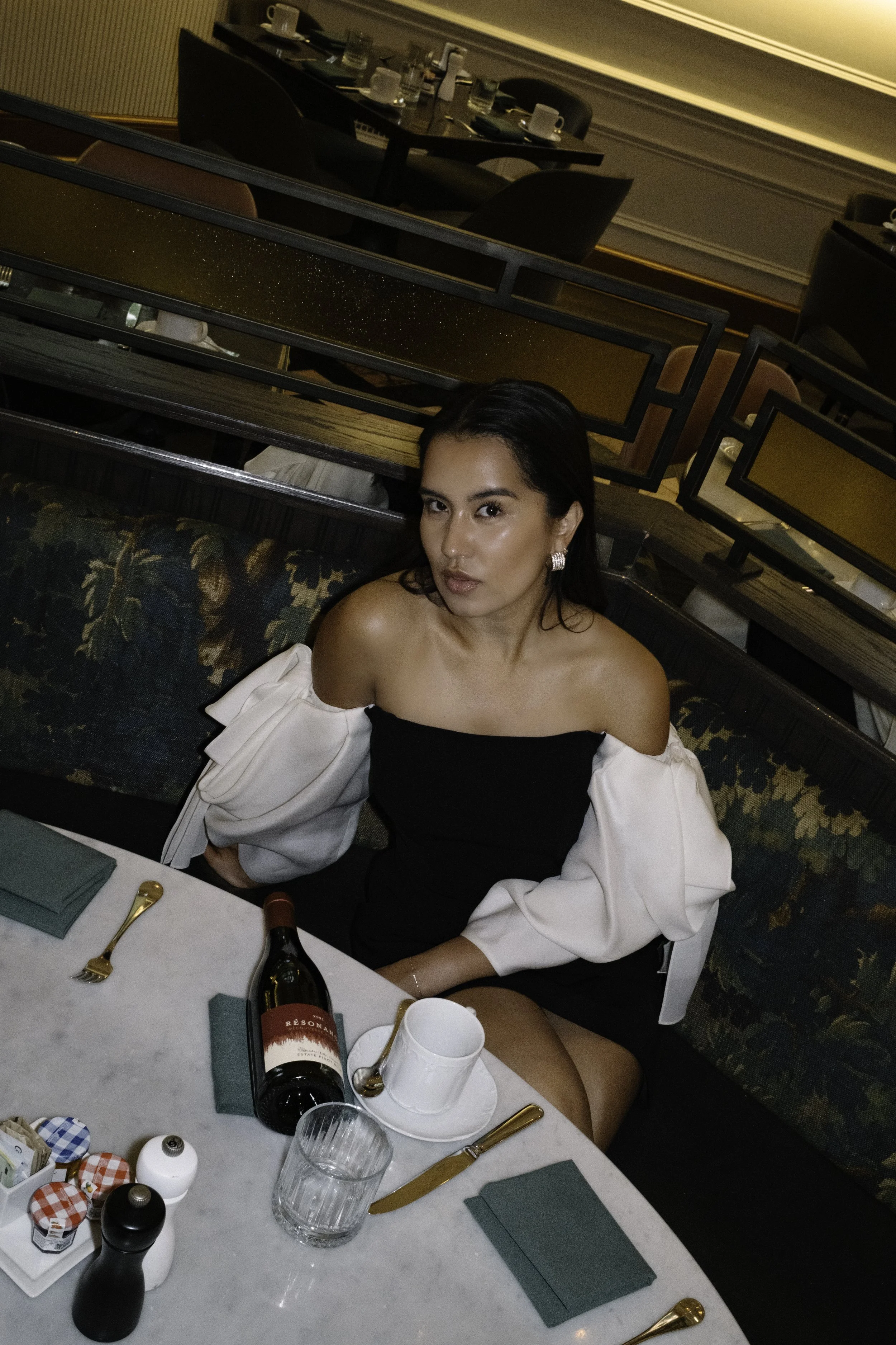 A woman sitting at a restaurant table with a white marble top, wearing a black and white off-shoulder dress, with a glass of water, a bottle of red wine, and some condiments on the table.