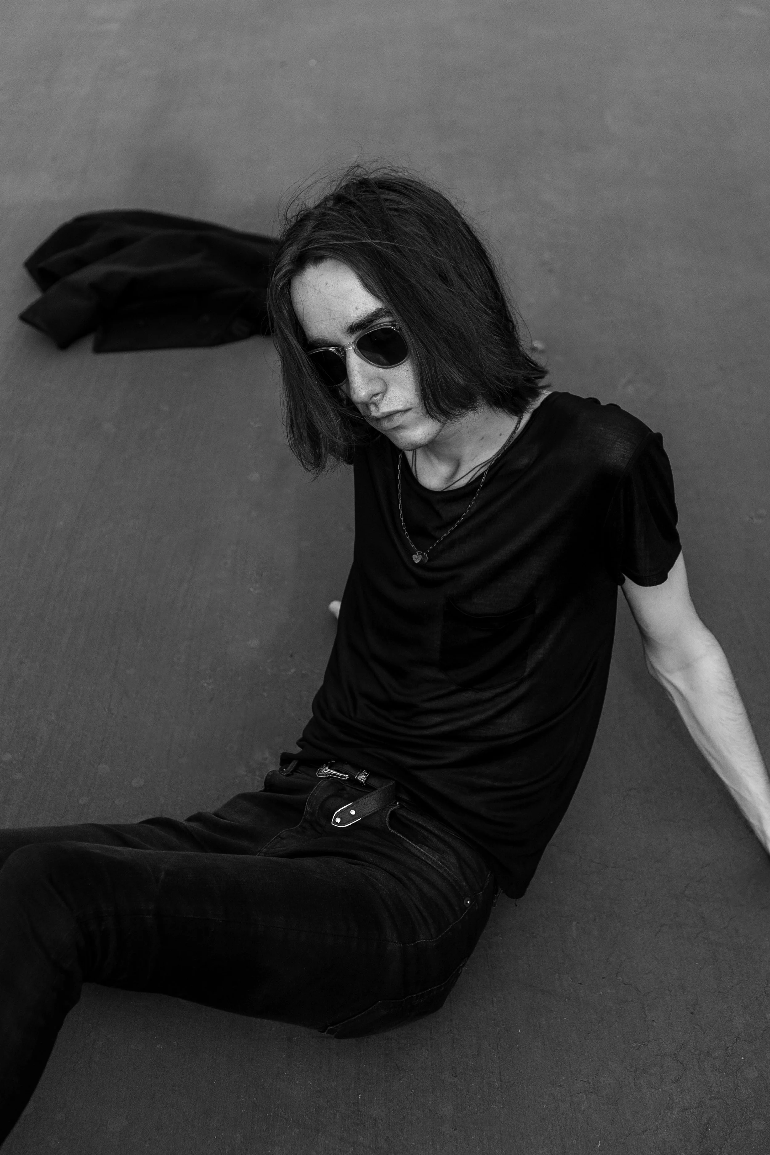 Black and white photo of a woman sitting on a textured surface, wearing sunglasses, a black t-shirt, black jeans, and a necklace, with a jacket lying behind her.