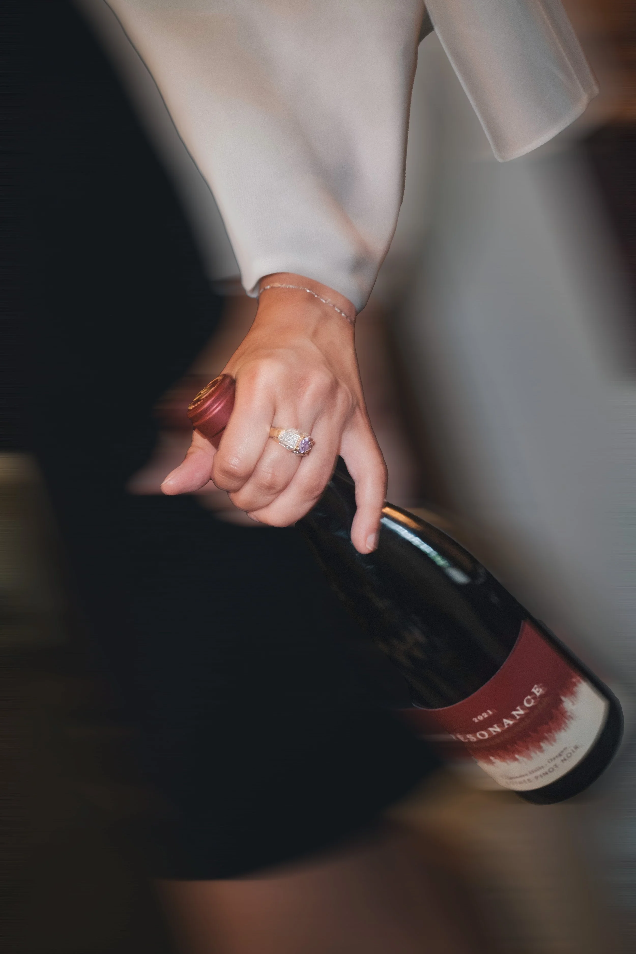 A woman's hand with jewelry, bending a wine bottle. She is wearing rings and a silver bracelet. She is dressed in a light-colored satin or silk garment.