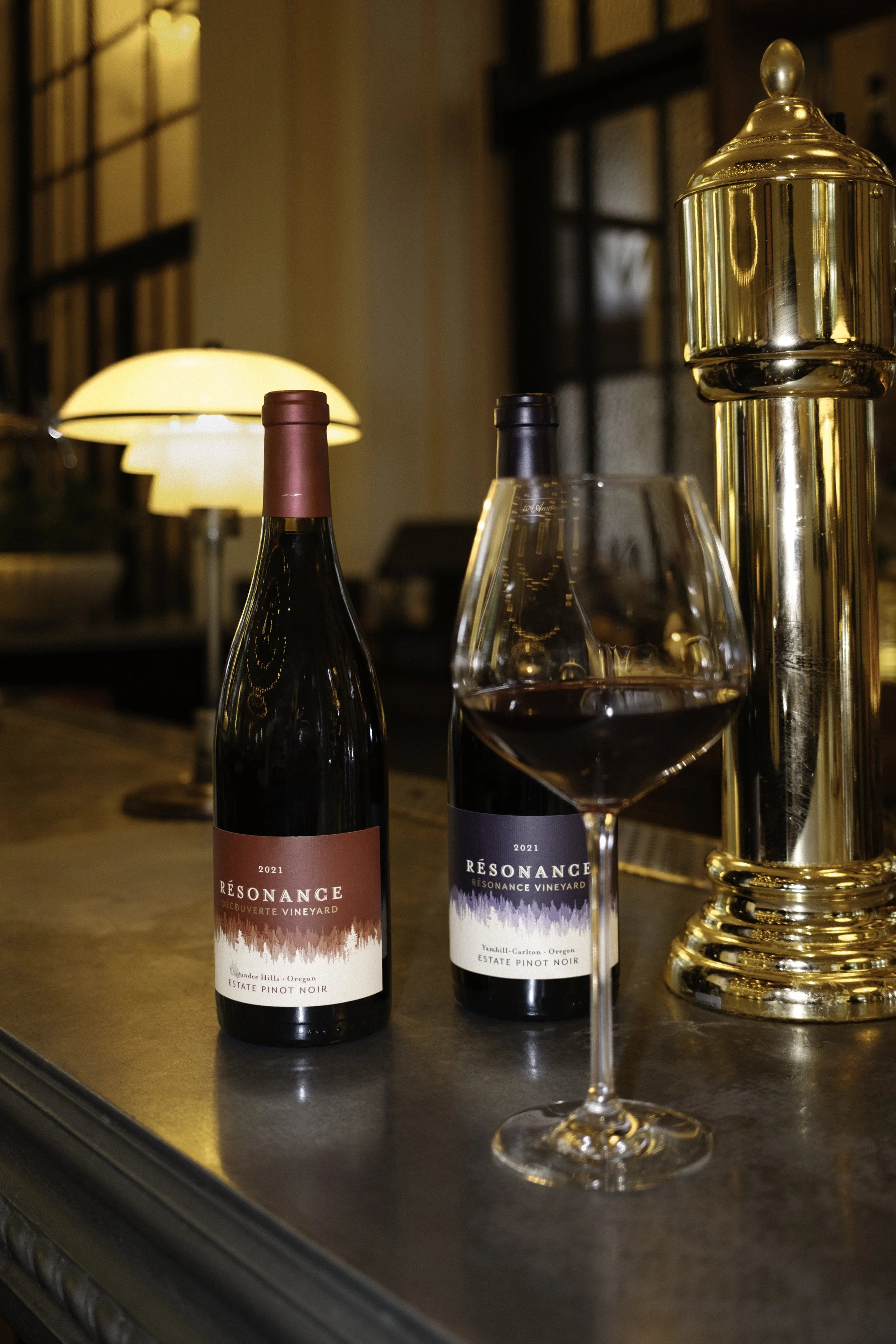 Two bottles of Resonance wine, one with a red label and the other with a black label, on a wooden bar counter with a glass of red wine in front of them. There is a gold-colored decorative object and a warm-lit lamp in the background.