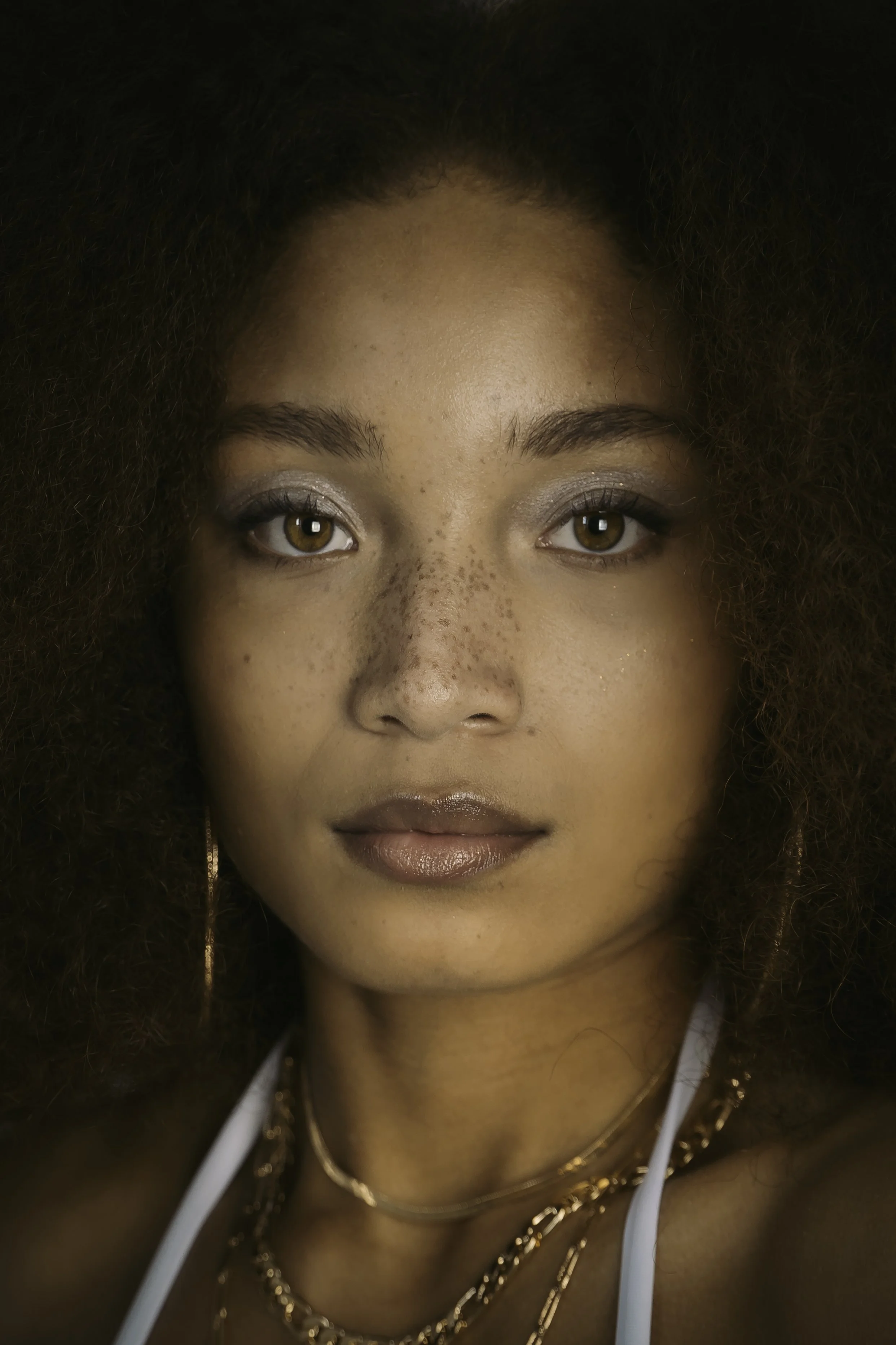 A close-up portrait of a woman with light brown skin, curly hair, light eye makeup, and gold jewelry.