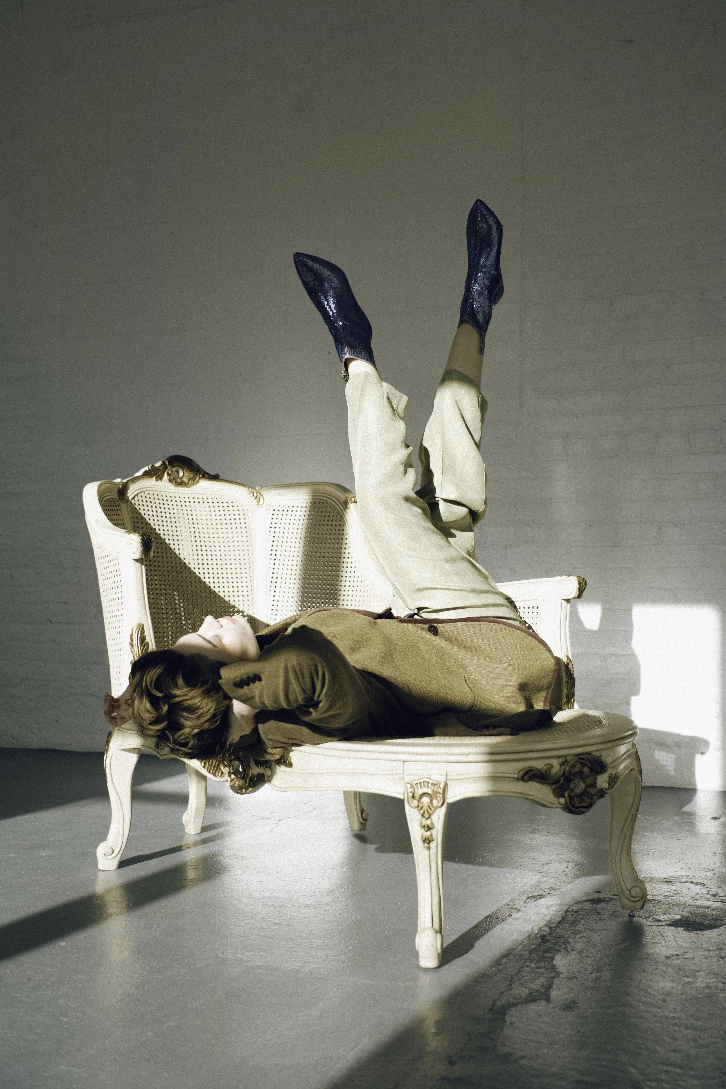 Person lying on an ornate vintage cream-colored sofa with legs up in the air, wearing beige pants, a brown blazer, and shiny dark blue boots, in a bright, minimalist room with white brick wall.