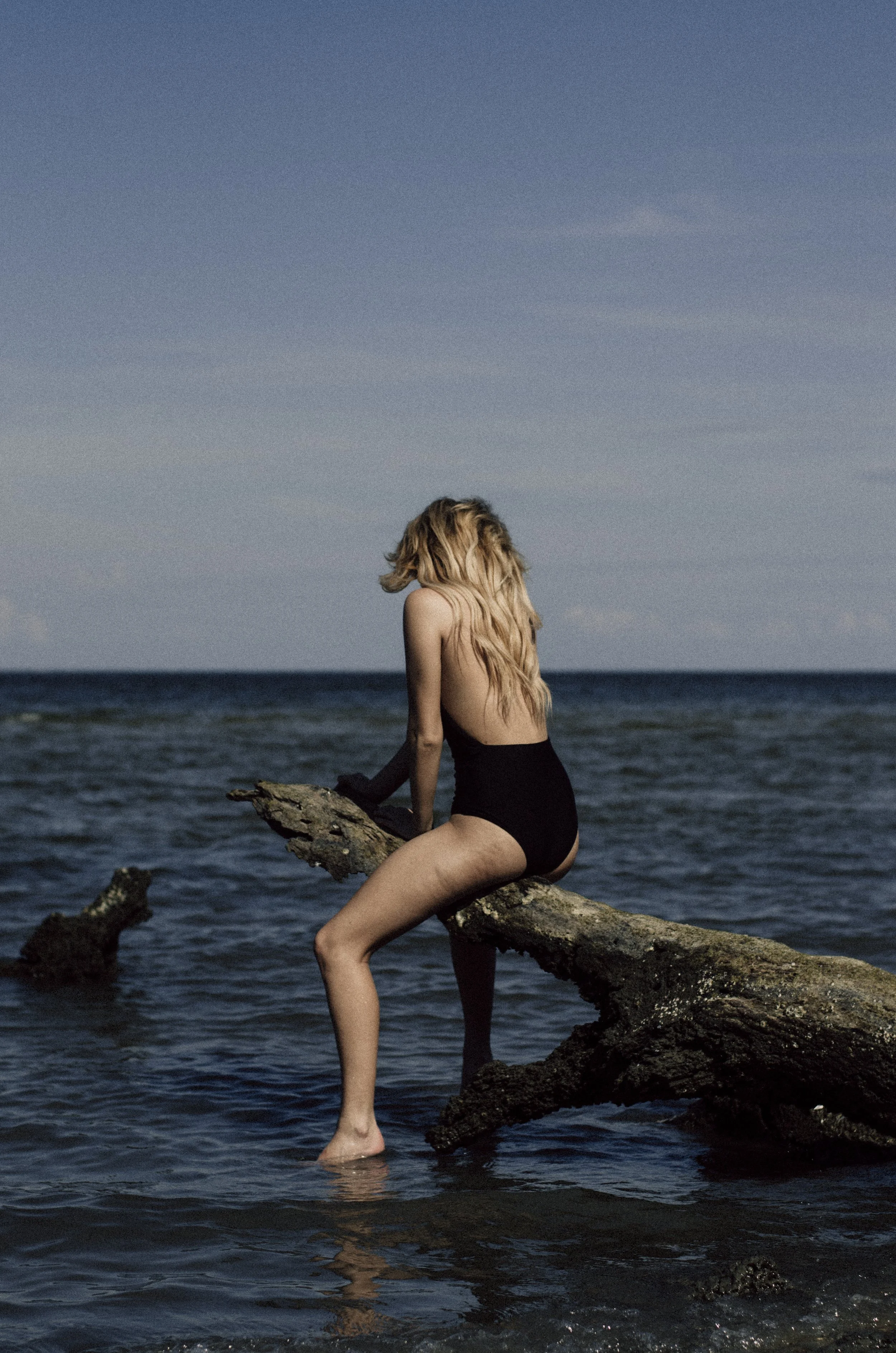 A woman with long blonde hair sitting on a large piece of driftwood in the water at the beach, wearing black high-waisted bikini bottoms and a black glove, with her back facing the camera and her head turned downward.
