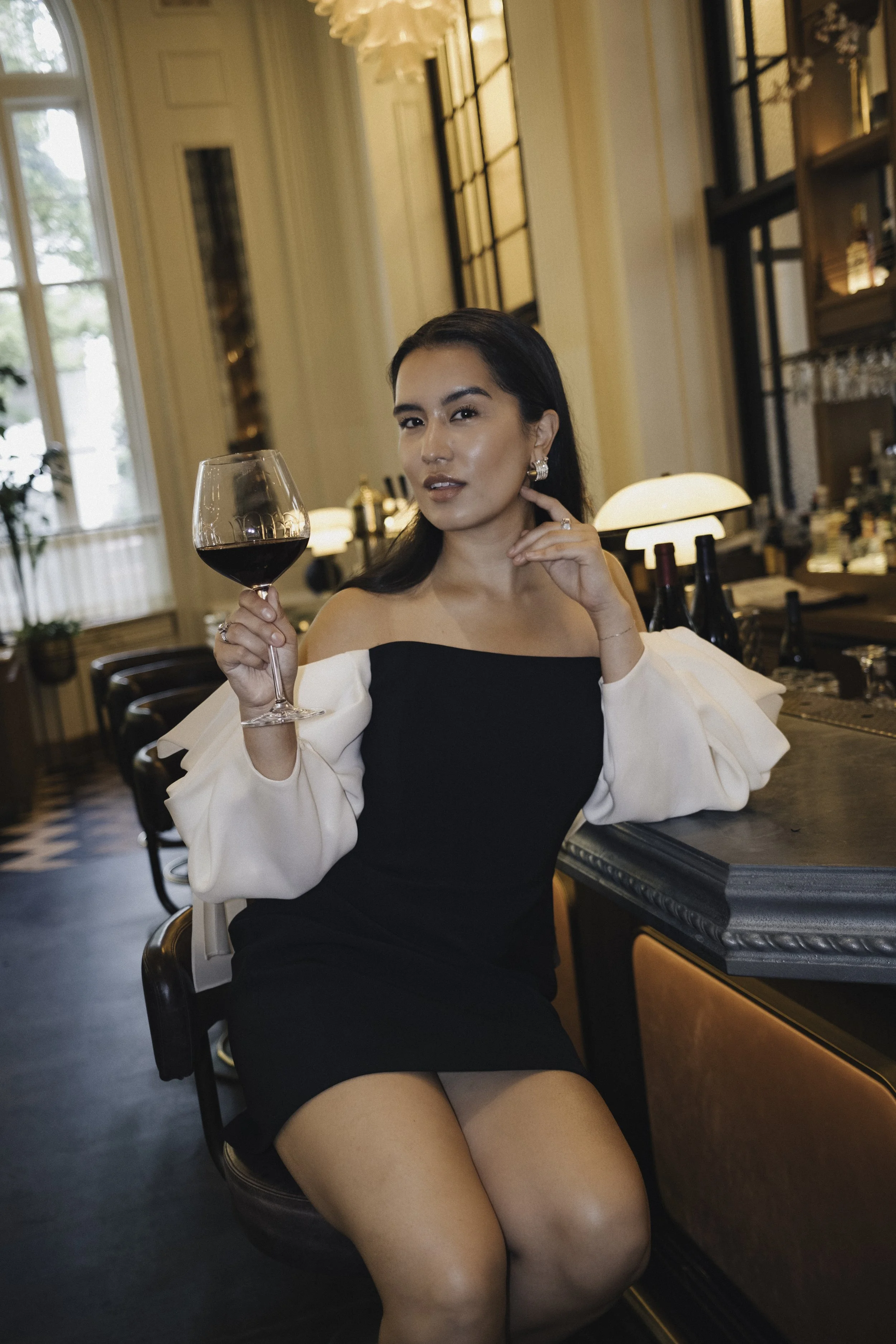 A woman in a black dress with white puffed sleeves sitting at a bar, holding a glass of red wine, in an elegant restaurant or bar setting with large windows and warm lighting.