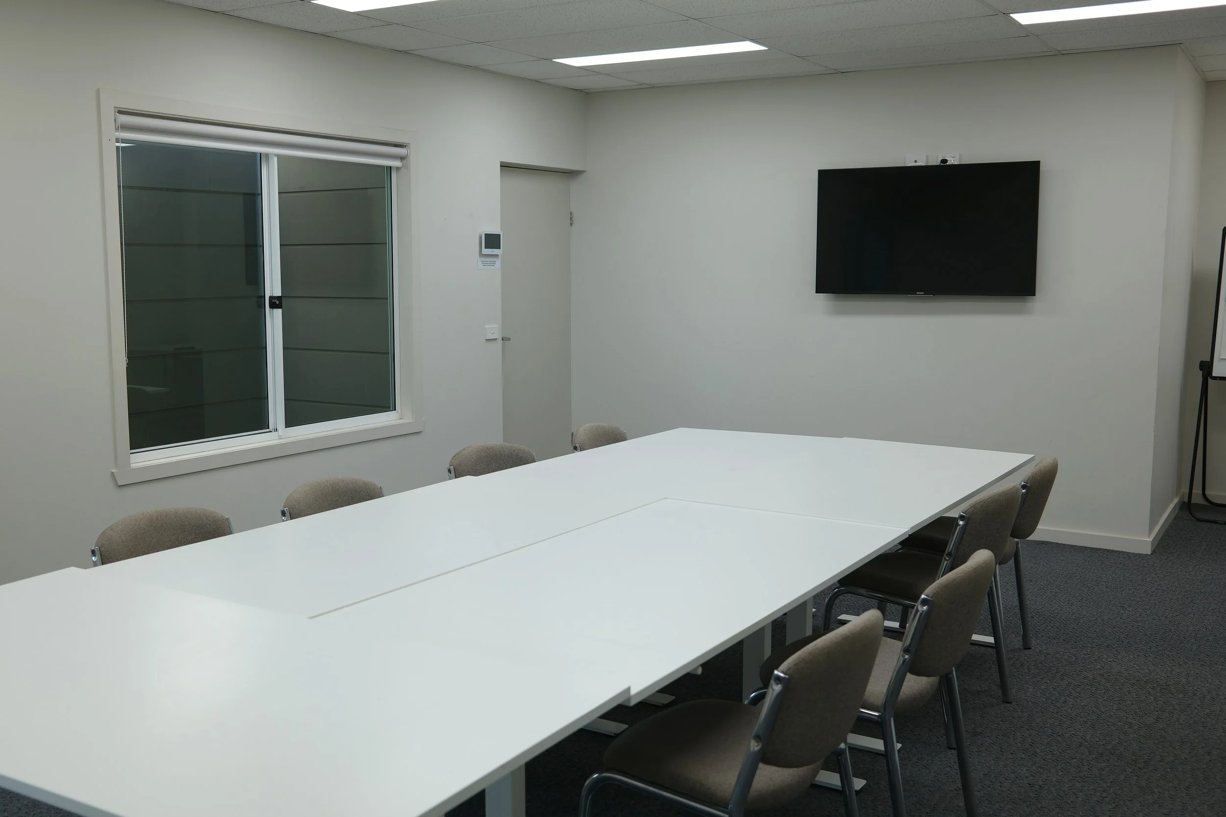 A conference room with a long white table and beige chairs. There is a large flat-screen TV mounted on the white wall, a sliding window with blinds, and a small thermostat or control panel beside the window. The room has a ceiling with fluorescent lighting and gray carpeted flooring.