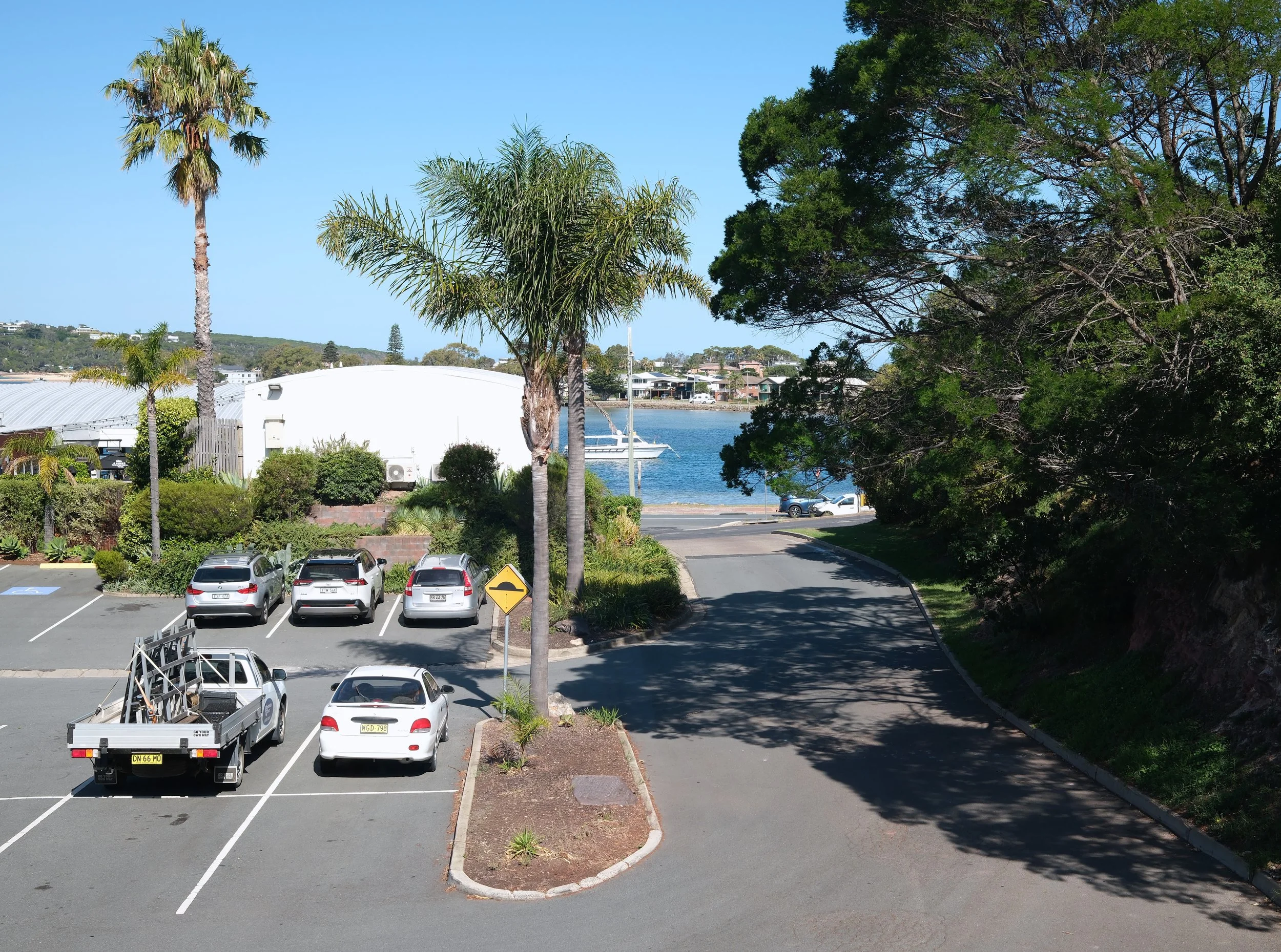 A parking lot next to a body of water with a boat, surrounded by trees and buildings, under a clear blue sky.