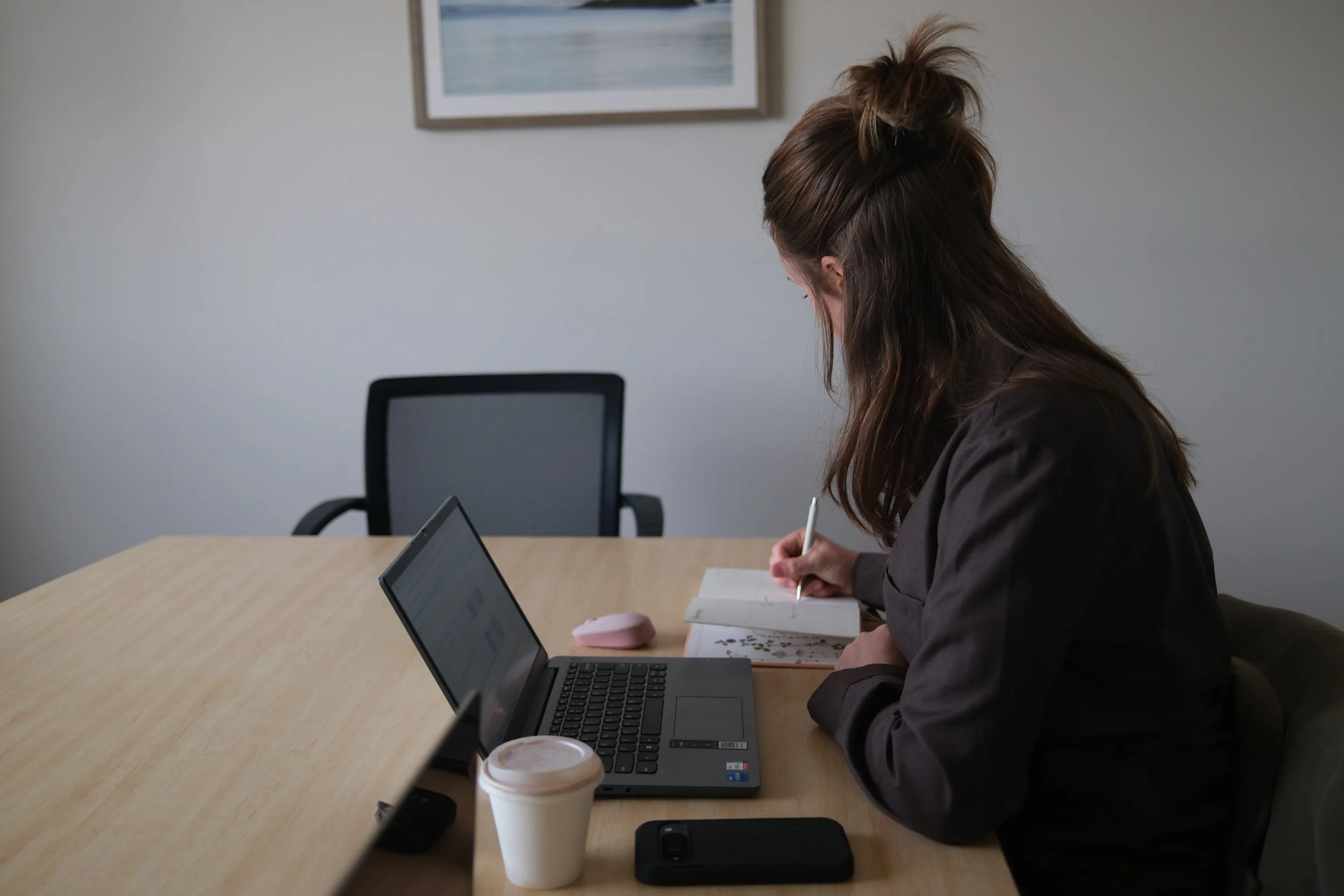 A woman with long hair tied up in a bun, sitting at a conference table, writing in a notebook, with a laptop, coffee cup, and smartphone in front of her, in a room with neutral walls and a framed picture hanging.
