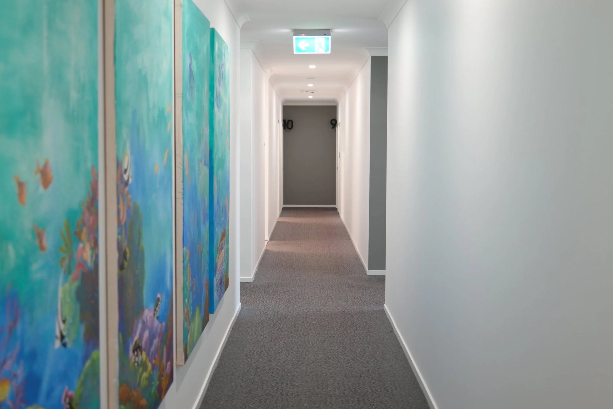 Interior hallway with white walls, featuring colorful abstract paintings on the left and a grey door numbered 9 at the end. There is an emergency exit sign hanging from the ceiling.