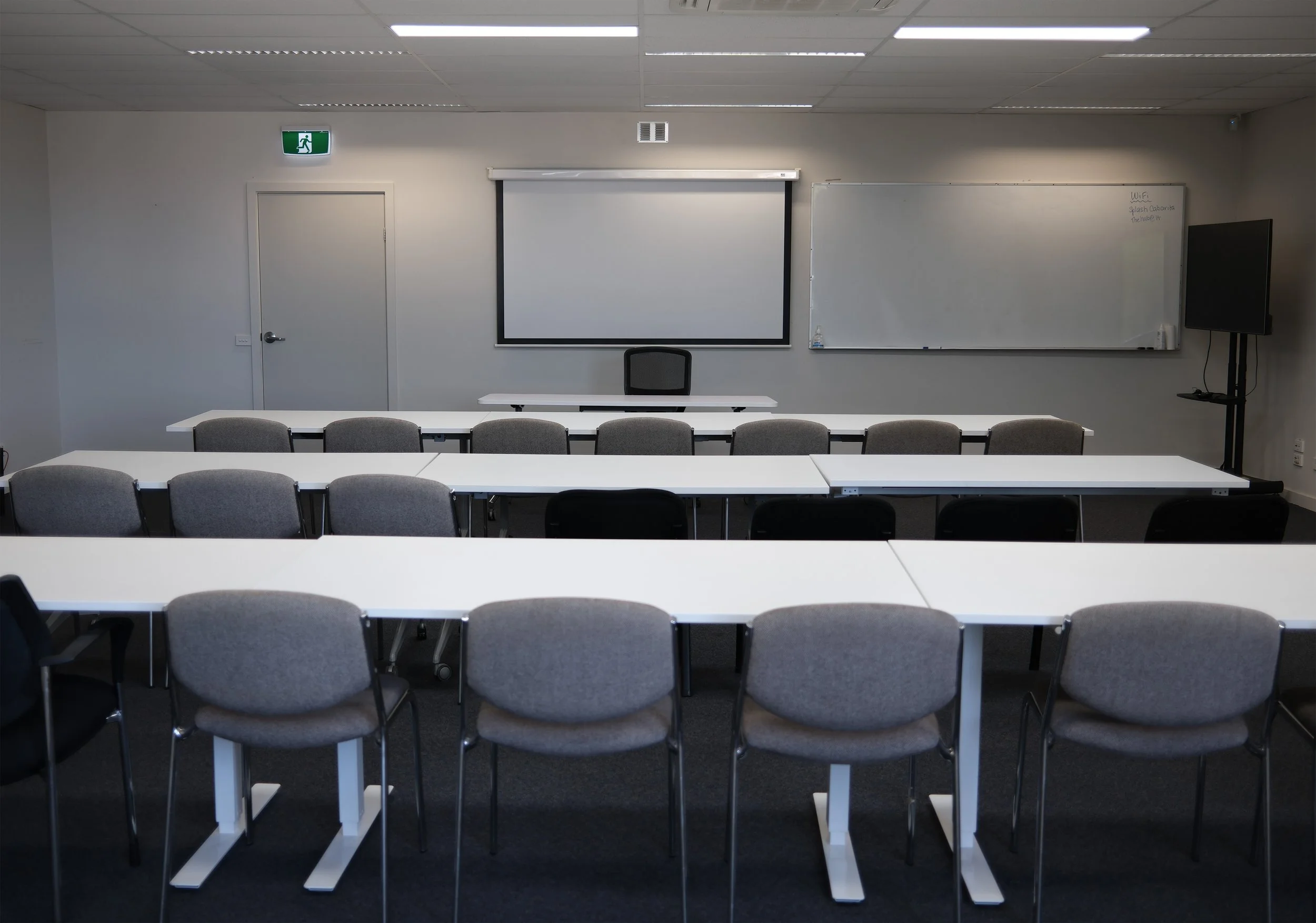 Empty classroom with white walls, ceiling lights, a whiteboard, a large monitor on a stand, and rows of desks and chairs.
