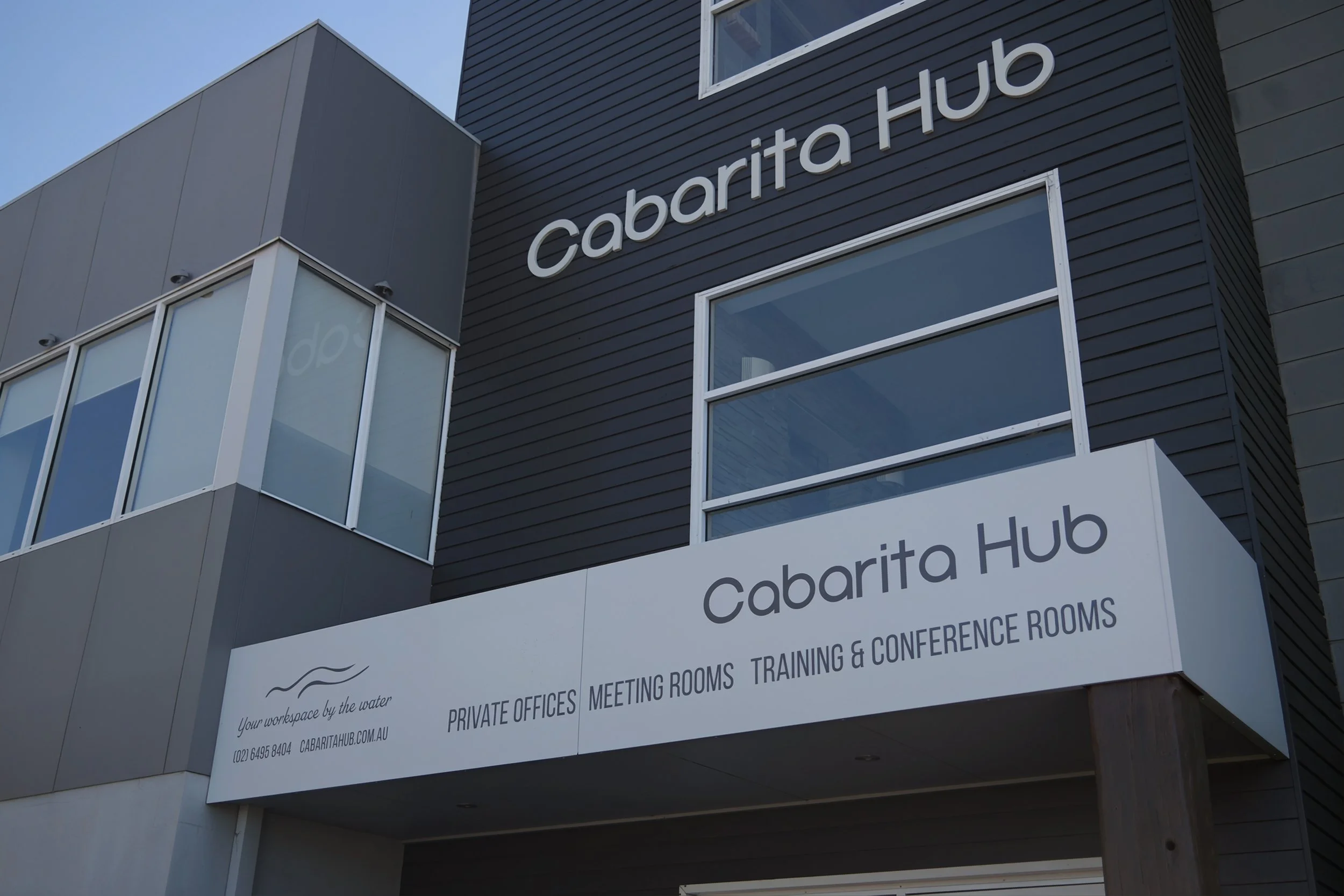 Exterior view of a modern multi-story building with black and gray siding, large windows, and a white sign reading "Cabarita Hub" with additional information about meeting rooms, conference rooms, and private offices.