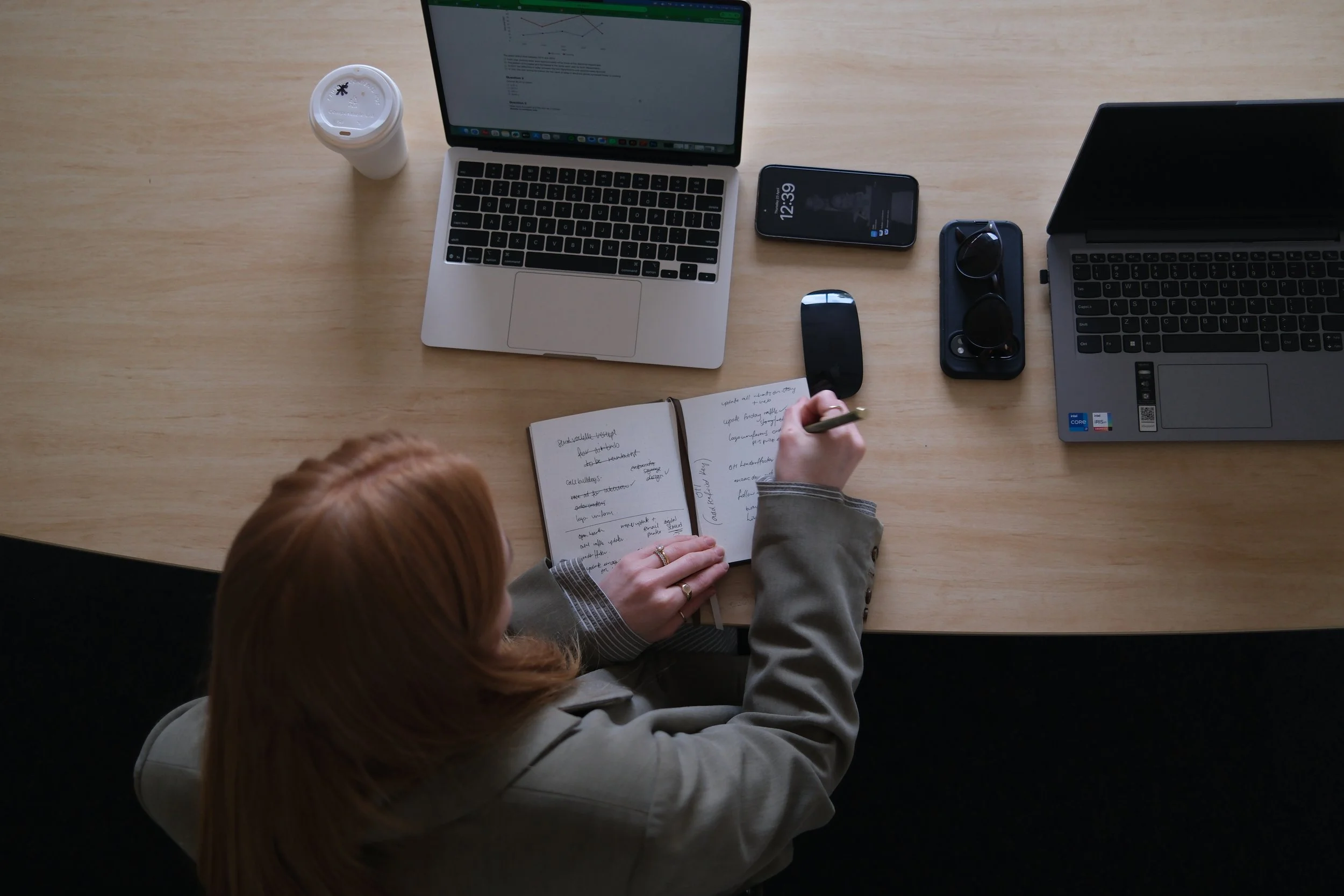 Top view of a workspace with a person writing in a notebook, surrounded by laptops, a smartphone, glasses, a mouse, a paper cup, and various documents on a wooden desk.