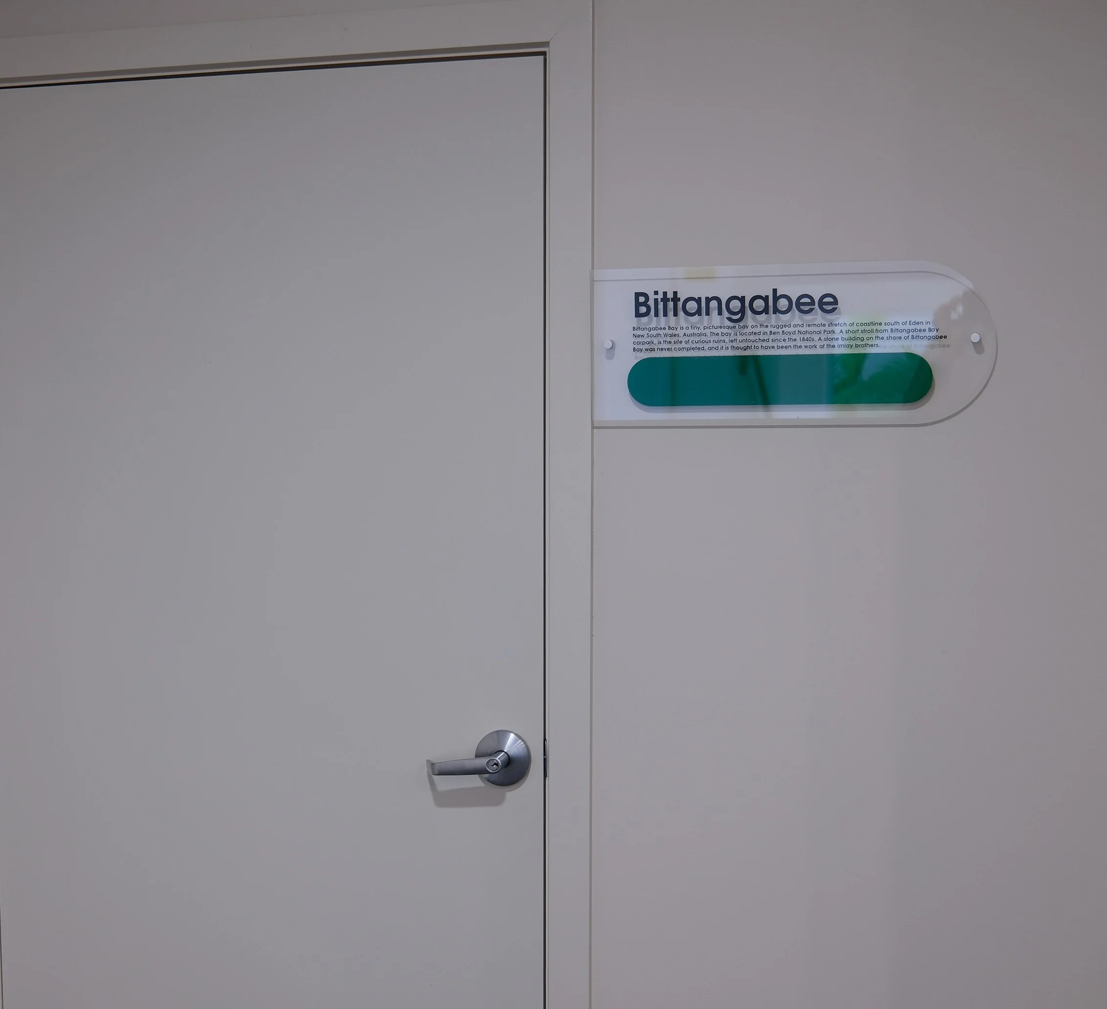 A gray door with a silver handle and a white informational sign attached to the upper part of the door. The sign contains text about Bittangabee Bay, located in South West Wales, Australia, and mentions its history and natural features.