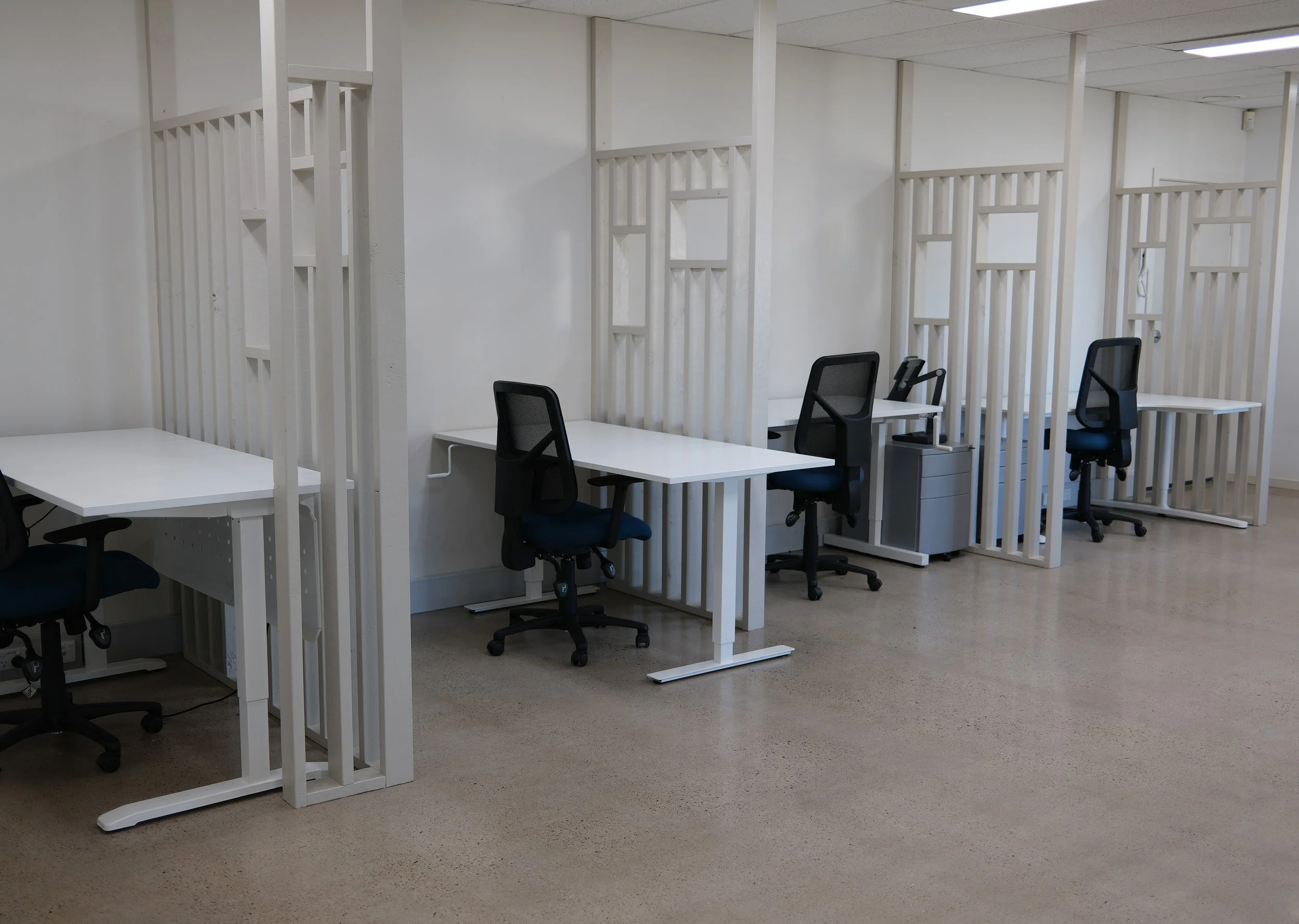 An office space with white desks separated by white frame partitions. There are black office chairs with mesh backs at each desk. The room has a white ceiling with fluorescent lighting and a plain beige floor.