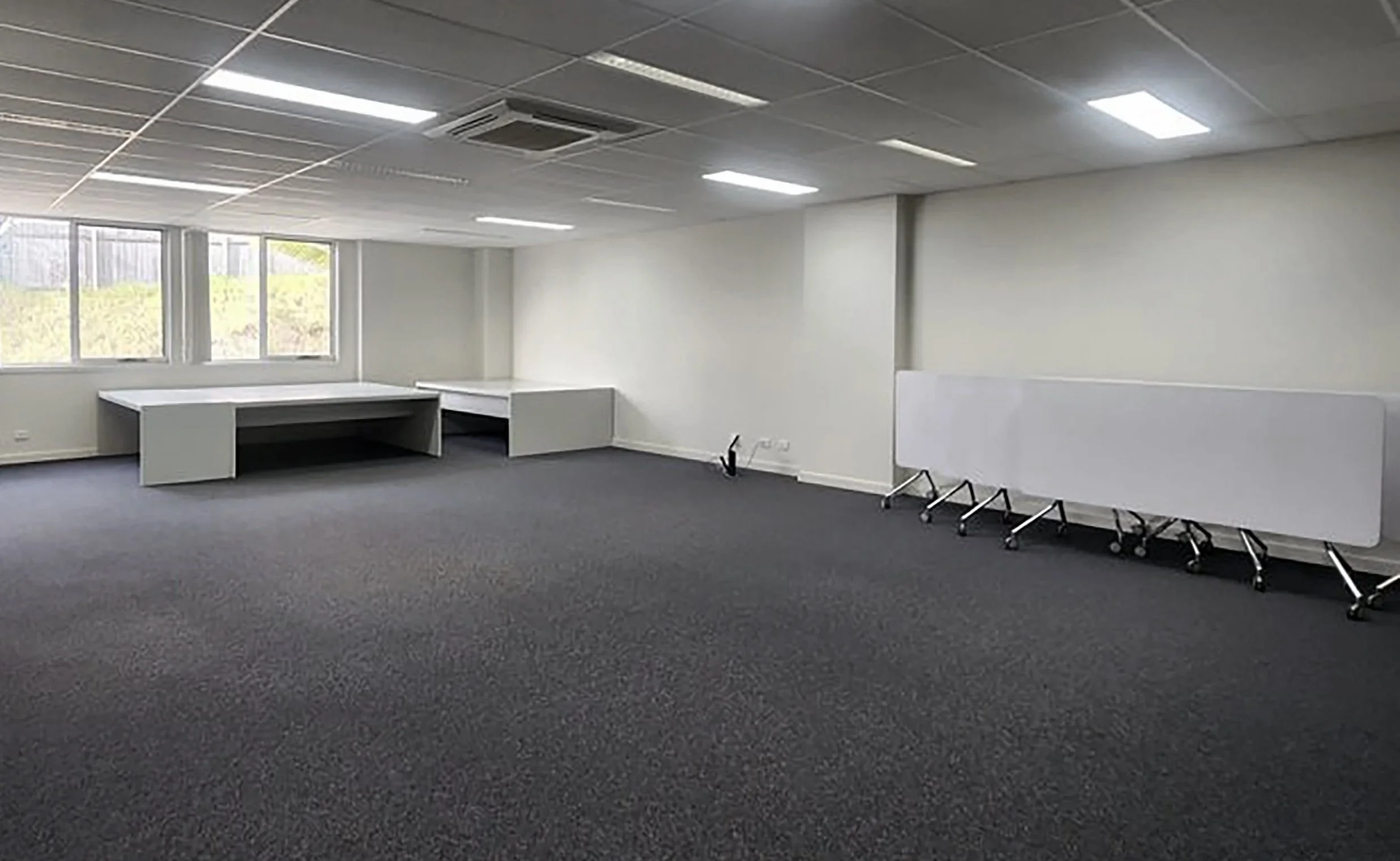 Empty office space with gray carpet, white walls, large windows, a white desk in the corner, and a whiteboard on wheels.
