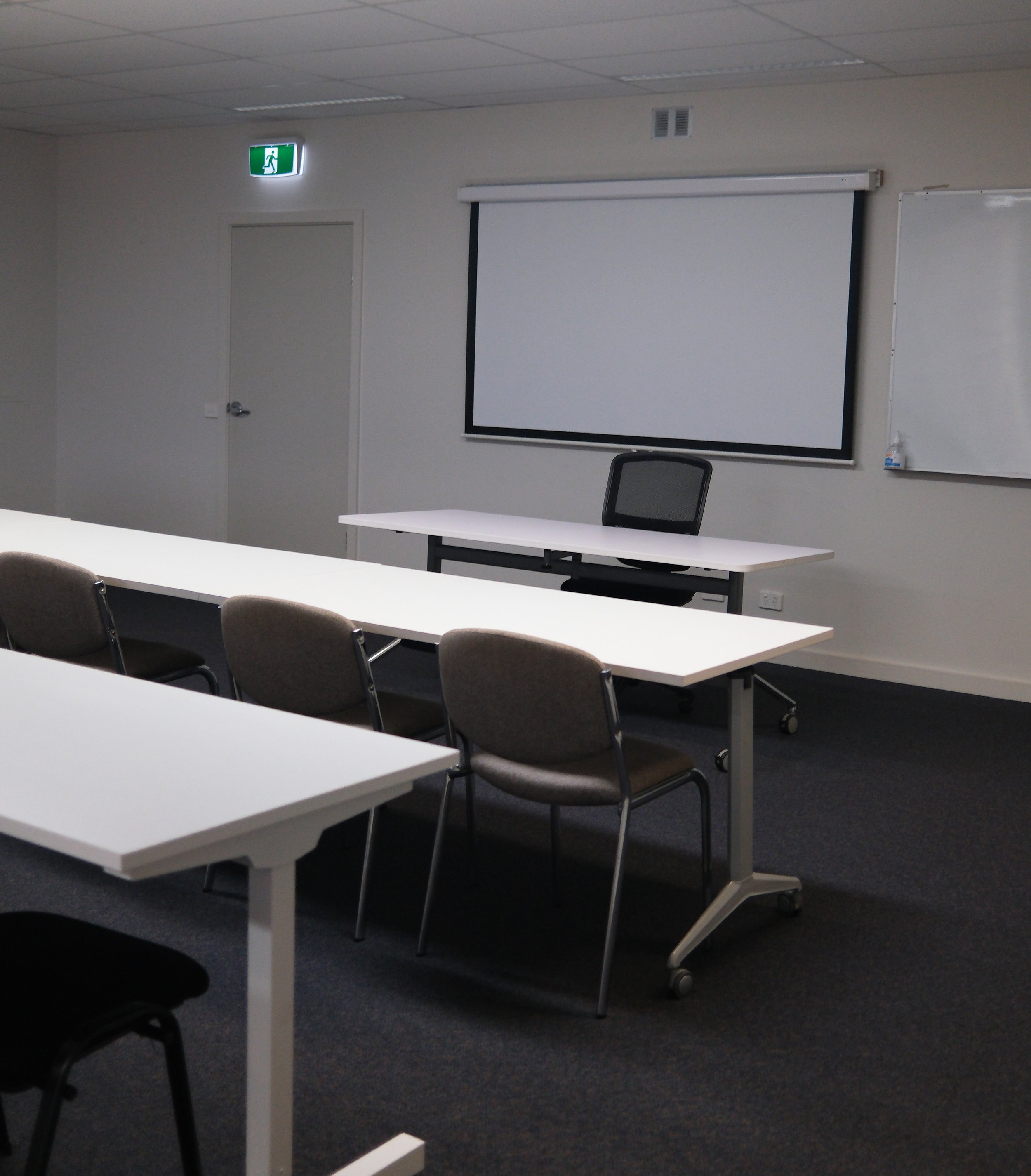 Empty classroom with white tables, gray chairs, a whiteboard, a projection screen, an exit sign, and a door.