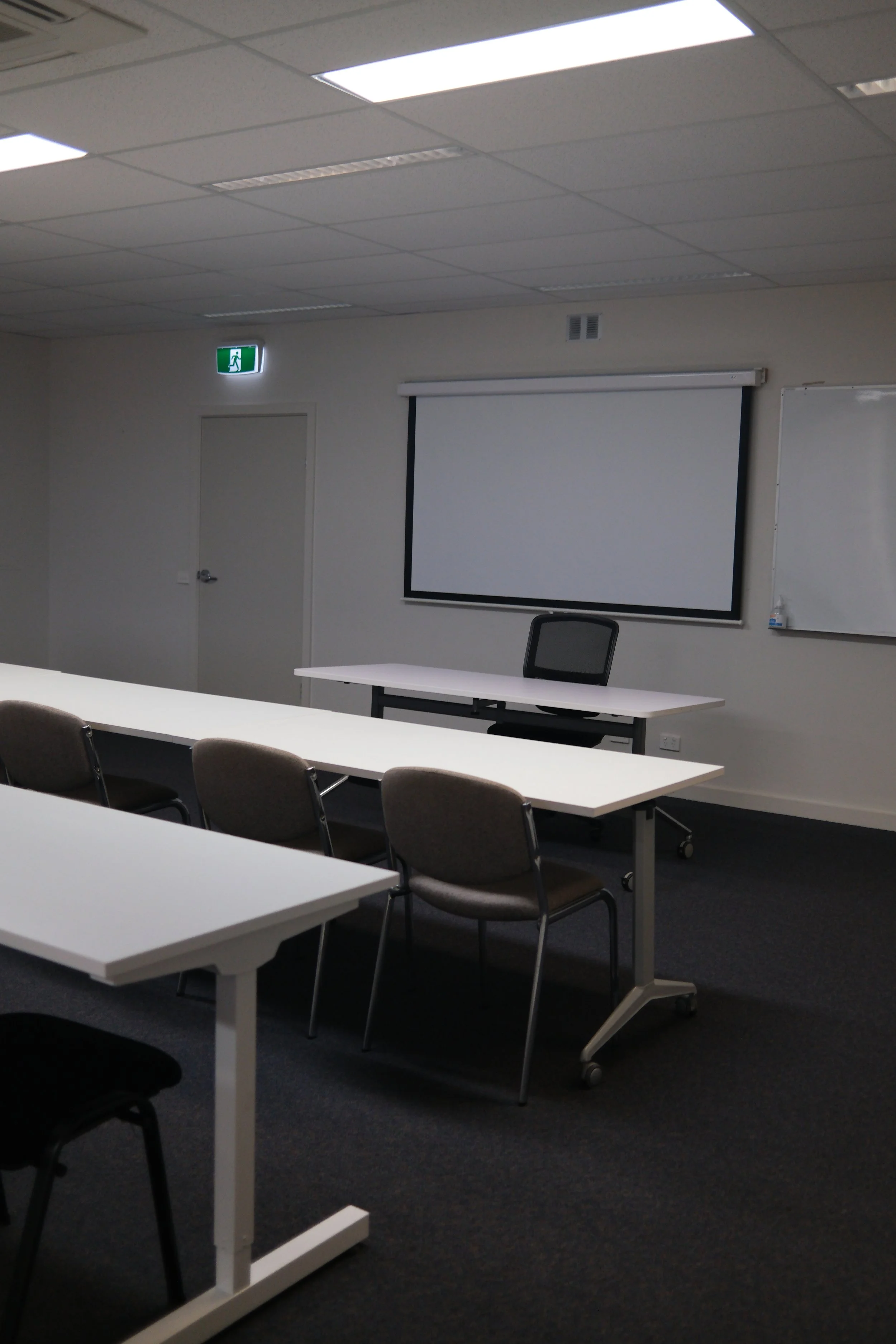 Empty meeting room with white tables arranged in a U-shape, chairs around them, a whiteboard on the right, a projection screen, a chair behind the main table, a ceiling with fluorescent lights, an exit sign above a door, and a dark carpeted floor.