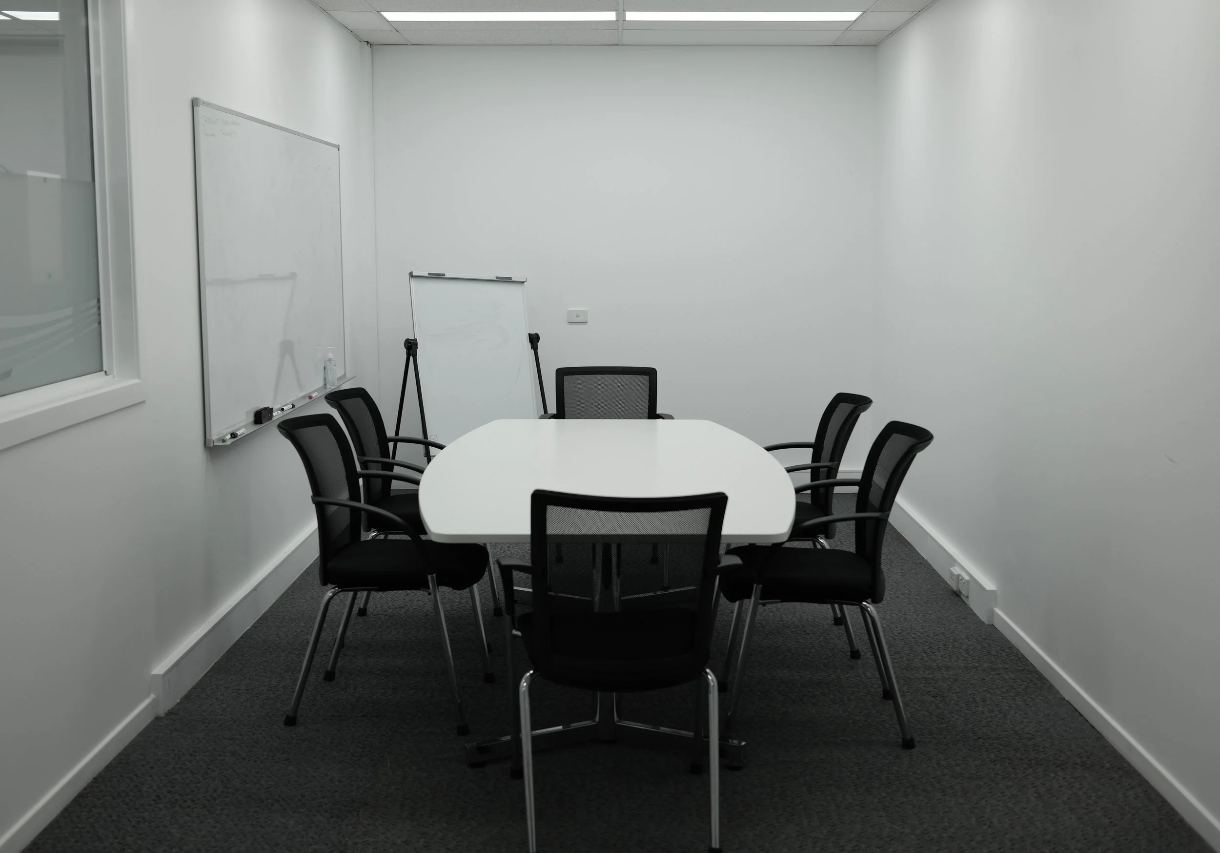 A small conference room with a white oval table and six black office chairs. There is a whiteboard on the wall, a flip chart on a stand, and a bottle of hand sanitizer on a shelf. The walls are white, and the flooring is dark gray carpet.