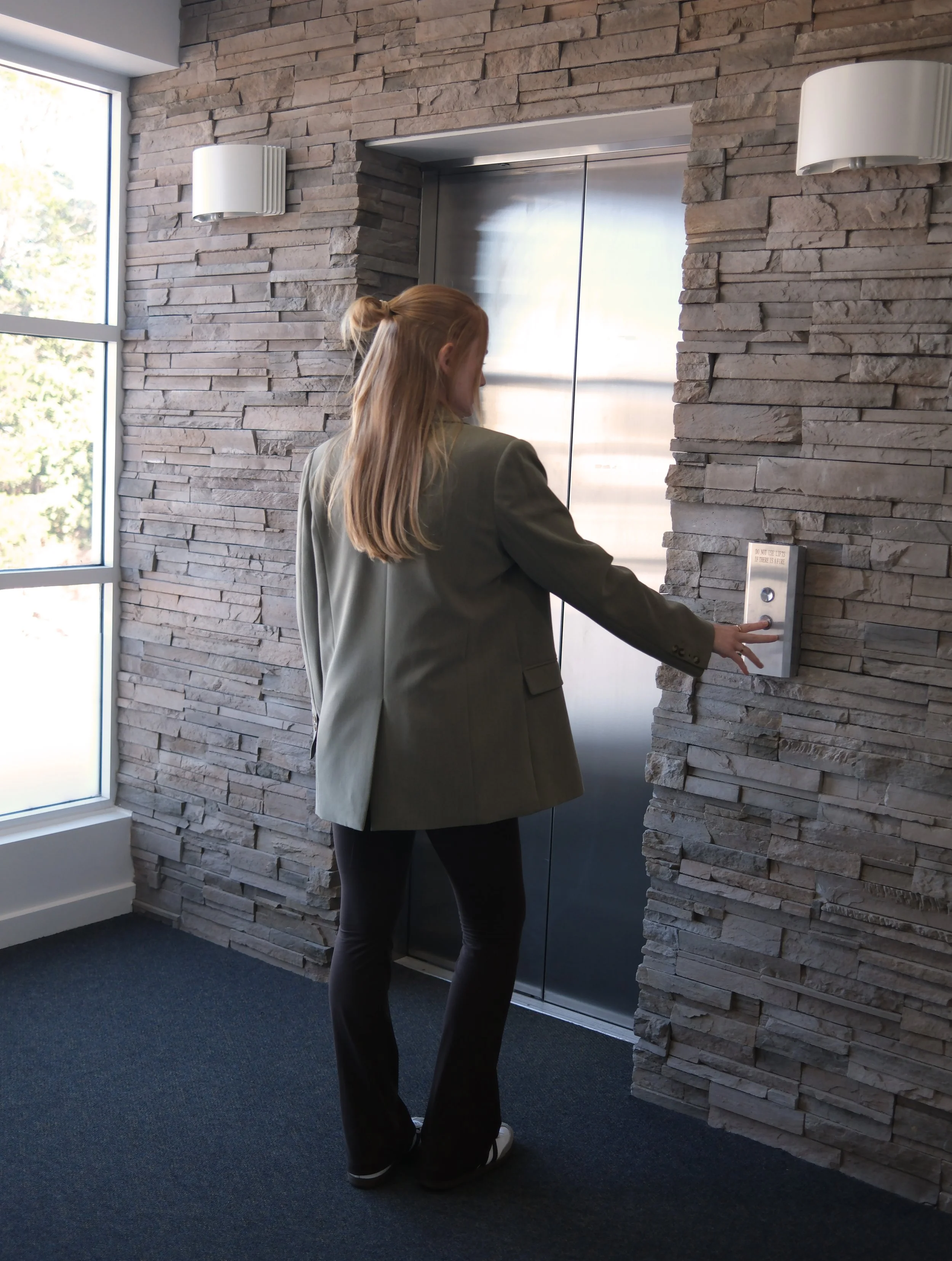 A woman with blonde hair, wearing a green blazer and black pants, is pressing the elevator button in a modern lobby area with stone walls and a large window.