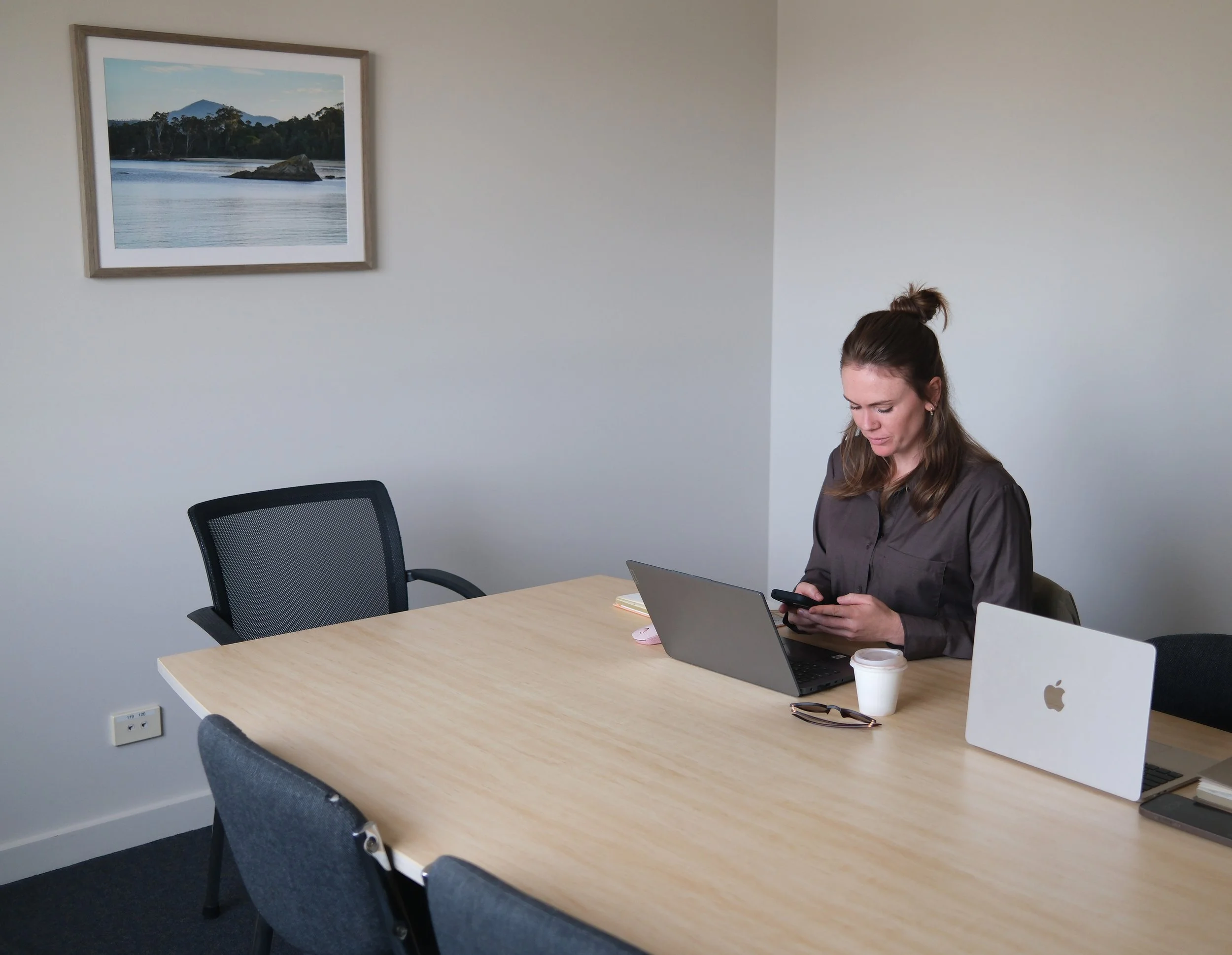 A woman sitting at a conference table in a meeting room, looking at her phone. The room has a framed landscape photograph of a lake and mountains on the wall. The table has a laptop, a paper coffee cup, and a desk phone. There are three empty chairs around the table.