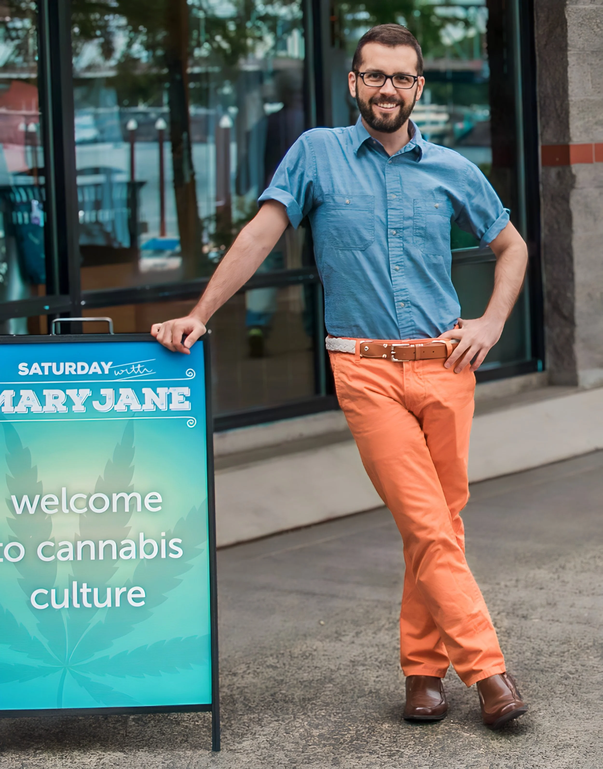 Alex Liberato standing next to a sign that says "welcome to cannabis culture" outside a storefront, wearing glasses, a blue shirt, orange pants, and brown shoes.