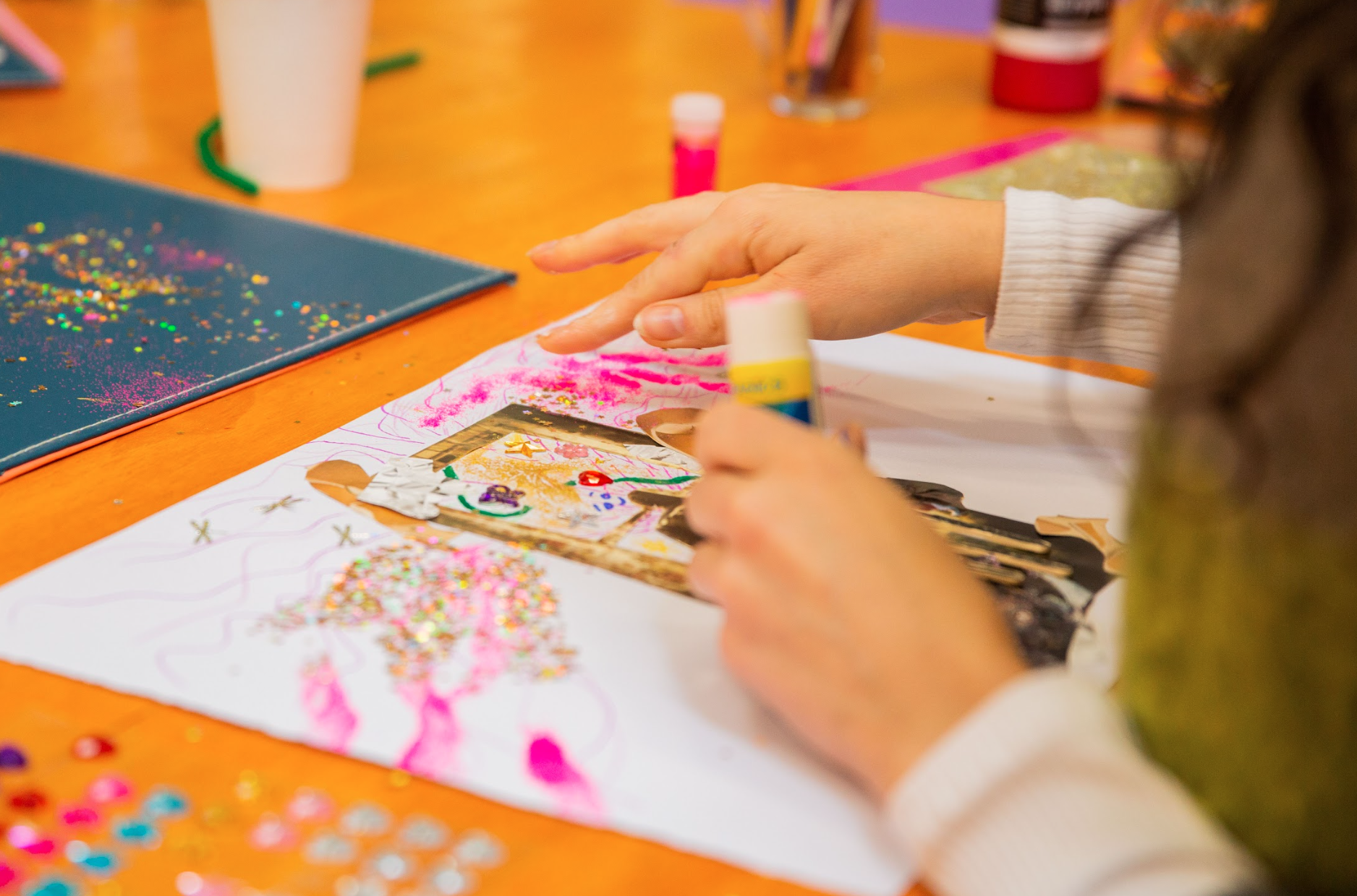 Person creating a collage with paper, glue stick, and glitter on a wooden table.
