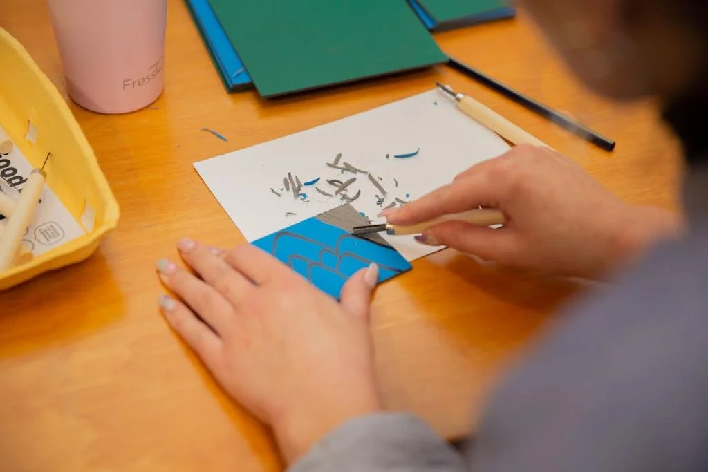 Person engaged in linocut art carving, with carving tools and linoleum block on a table.