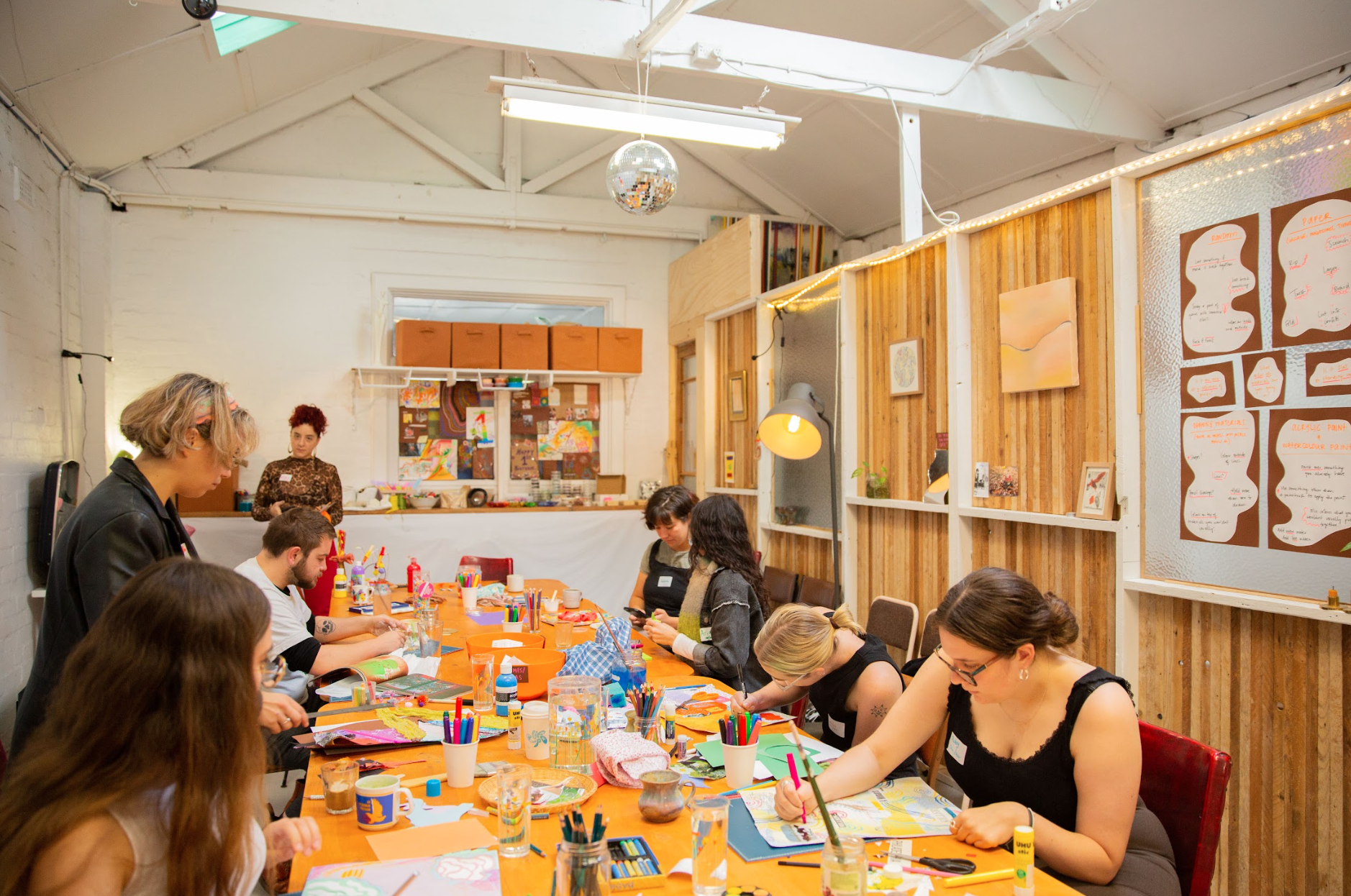 A group of people participating in an art workshop inside a creative studio. They are seated around a large table with various art supplies such as pencils, markers, and paper. The room has wooden paneling and artwork displayed on the walls. A person stands observing in the background. The atmosphere appears relaxed and collaborative.