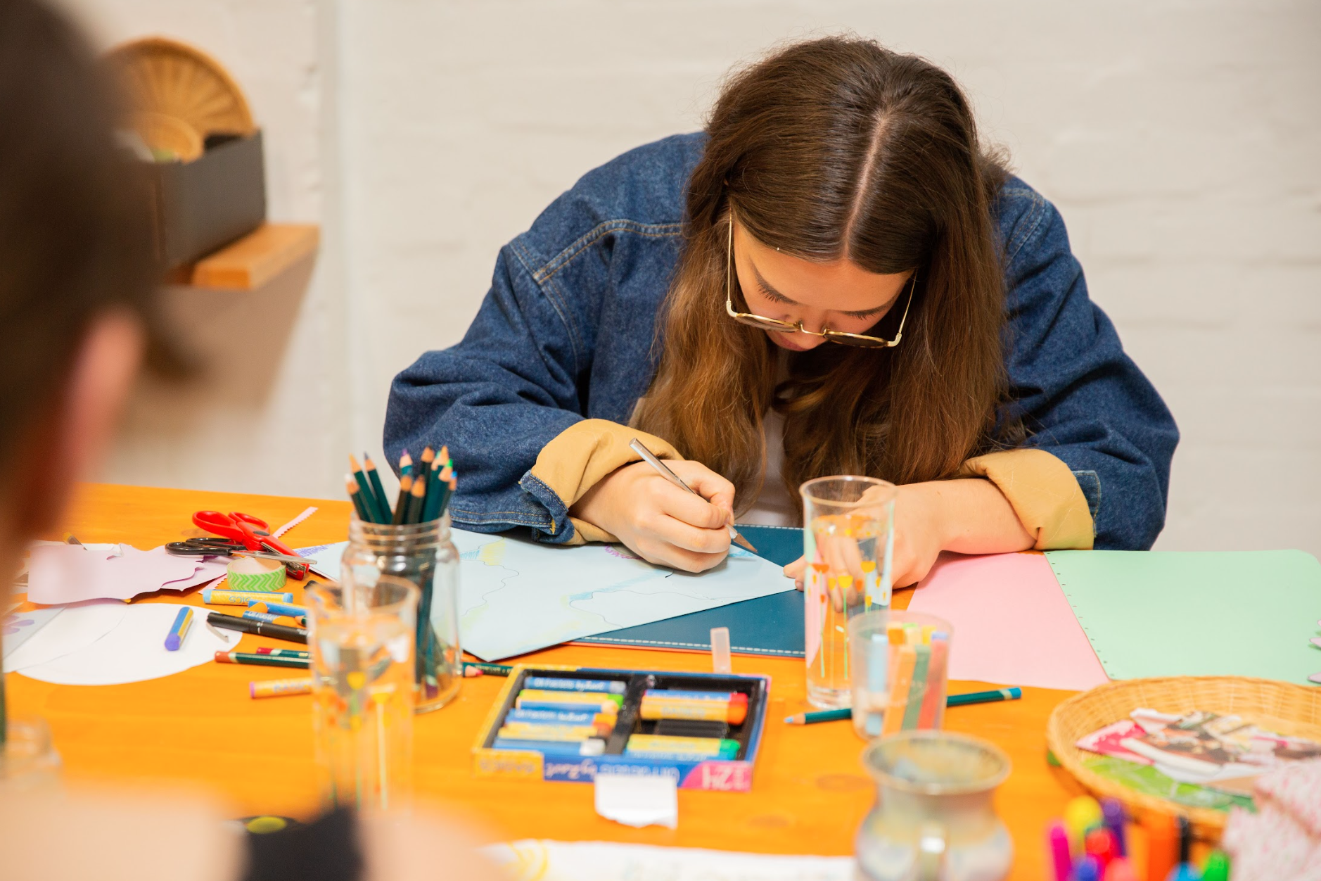 A person in a denim jacket is drawing or crafting at a table with various art supplies, including colored pencils, scissors, and glue sticks, on an orange tablecloth.