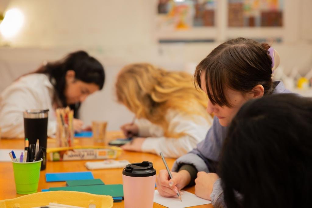 People sitting at a table writing on paper with pens and markers, surrounded by cups and stationery.