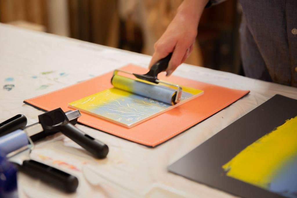 Person using a brayer to spread paint on a canvas or paper, with additional brayers and art materials on the table.