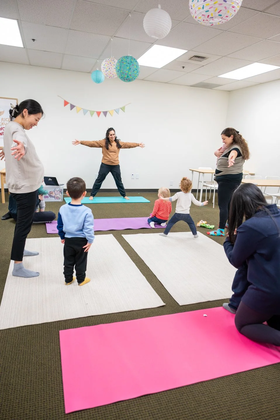A yoga instructor doing a yoga pose to show the toddlers some yoga moves while they all stand on yoga mats and area rugs.
