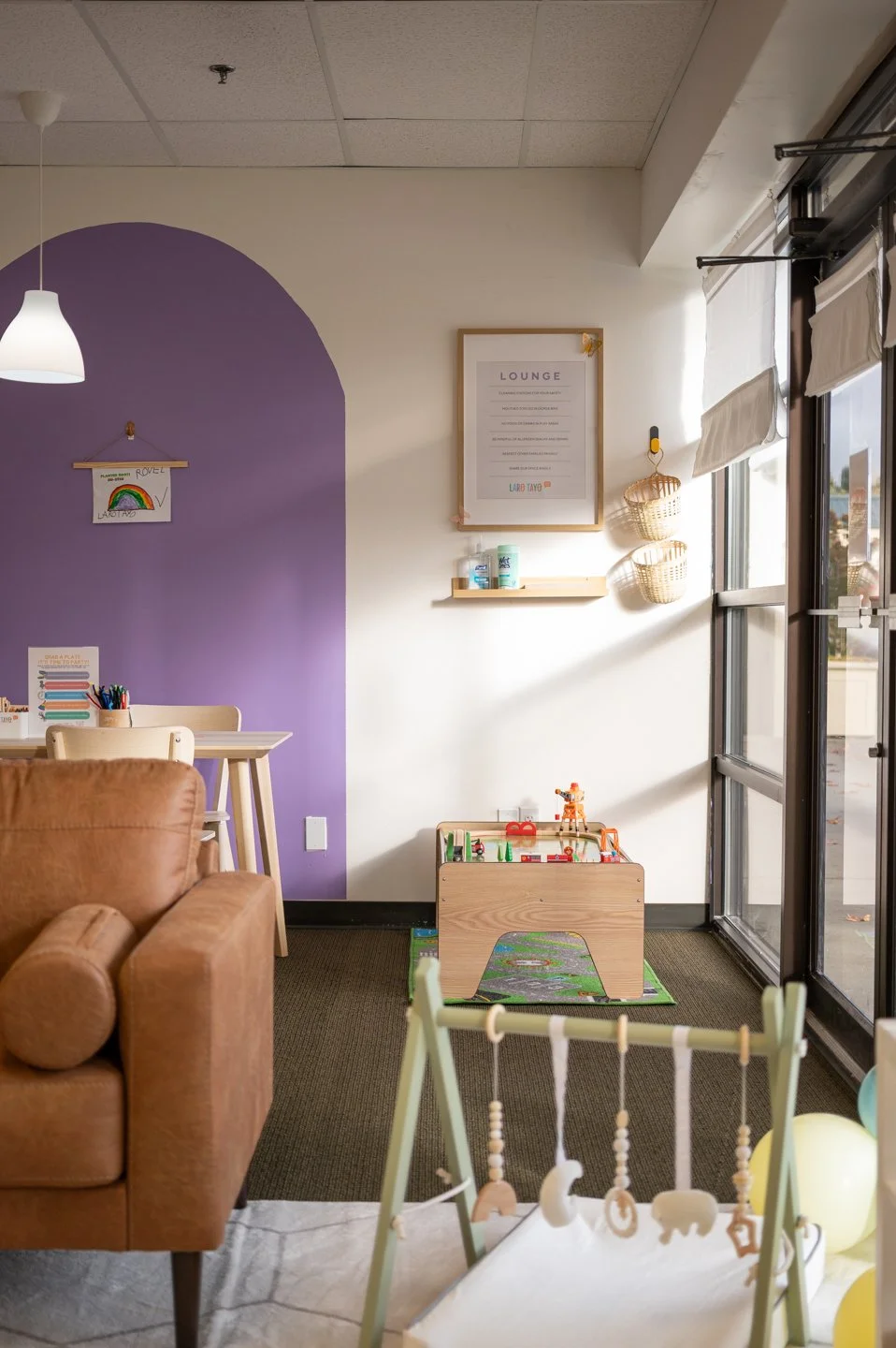 Inside of Laro Tayo by the emergency exit is the train table in the corner front wall, next to a wooden dining table with crafts on top, a leather couch in the foreground next to a baby pad with baby gym to encourage play for infants and babies.