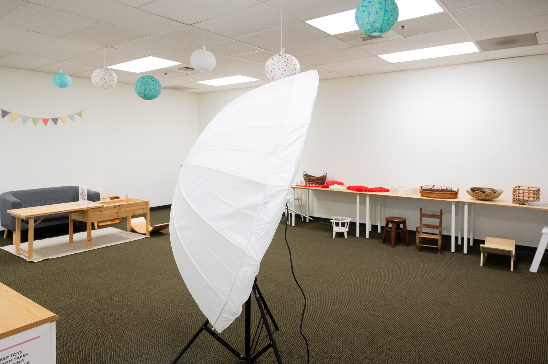 A big light umbrella on a tripod in the middle of the photo flanked by a couch and table and a table full of props for photo sessions